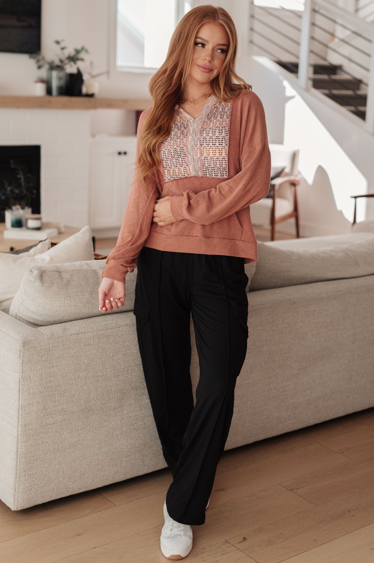 Woman modeling a rust hooded jersey top with a multicolored textured panel and black pants beside a sofa indoors