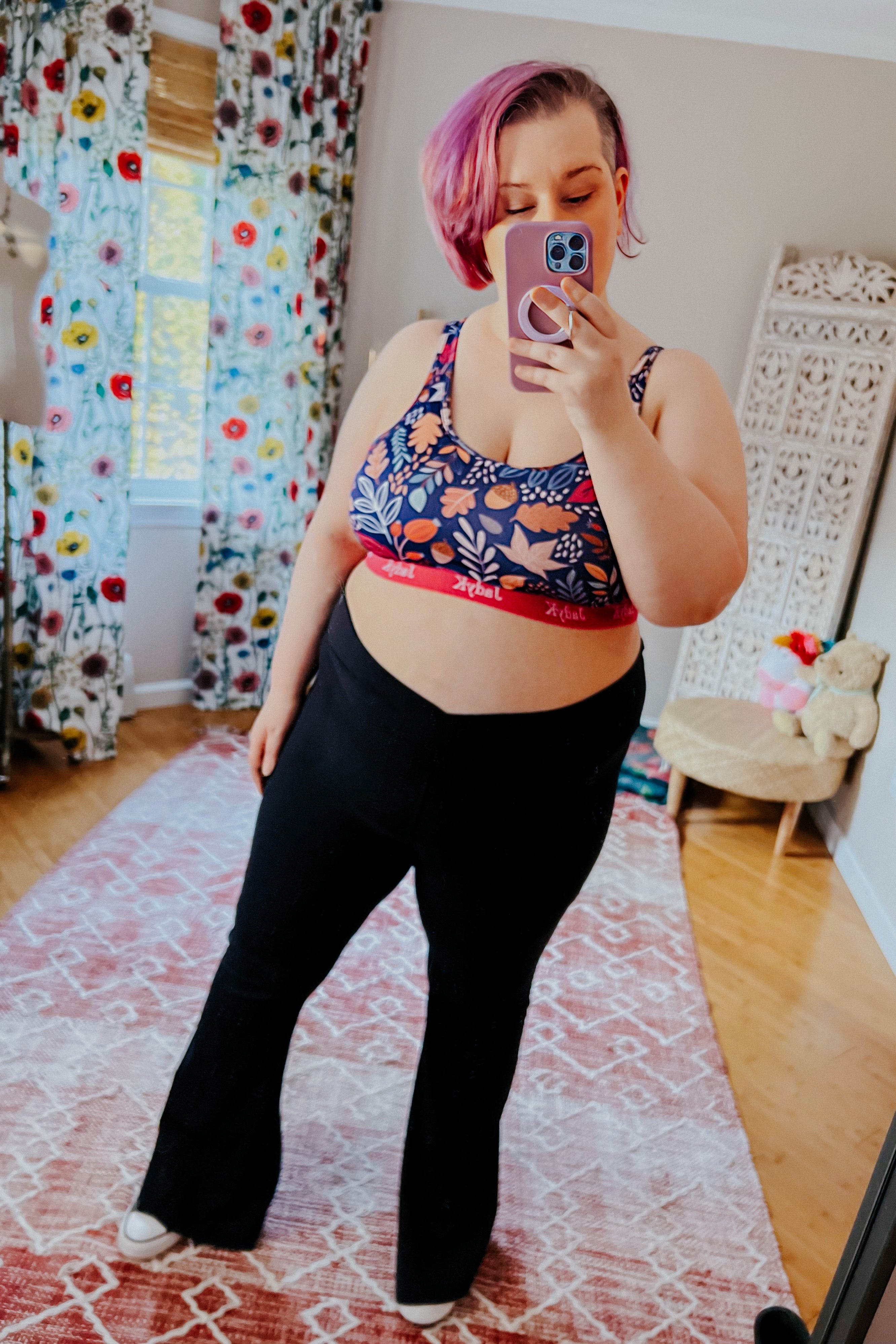 Plus-size woman with pink hair wearing a bold floral sports bra with red band and black pants taking a mirror selfie indoors