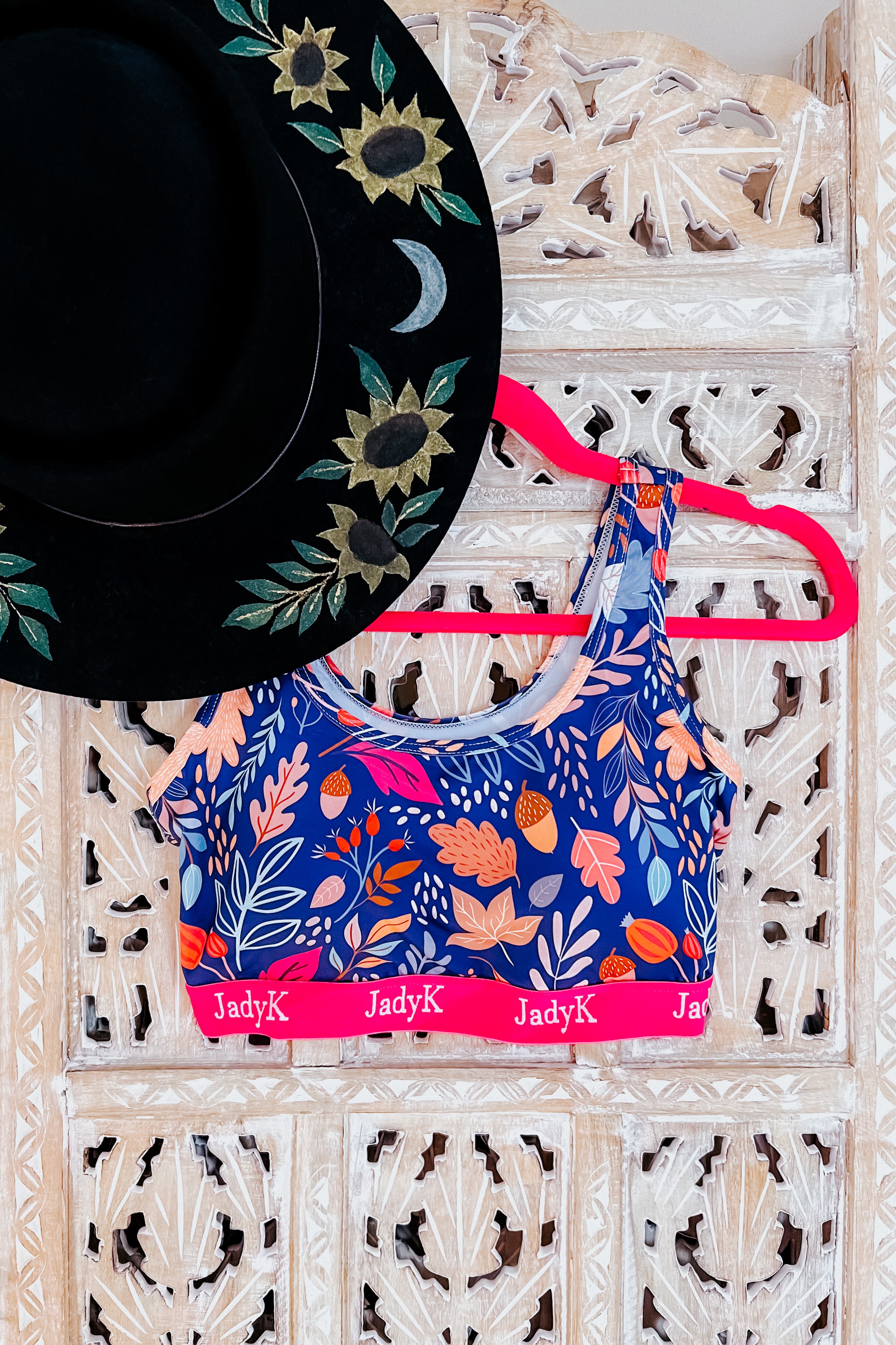 Blue printed bra with pink band and JadyK logo hanging on bright pink hanger next to black embroidered wide-brim hat