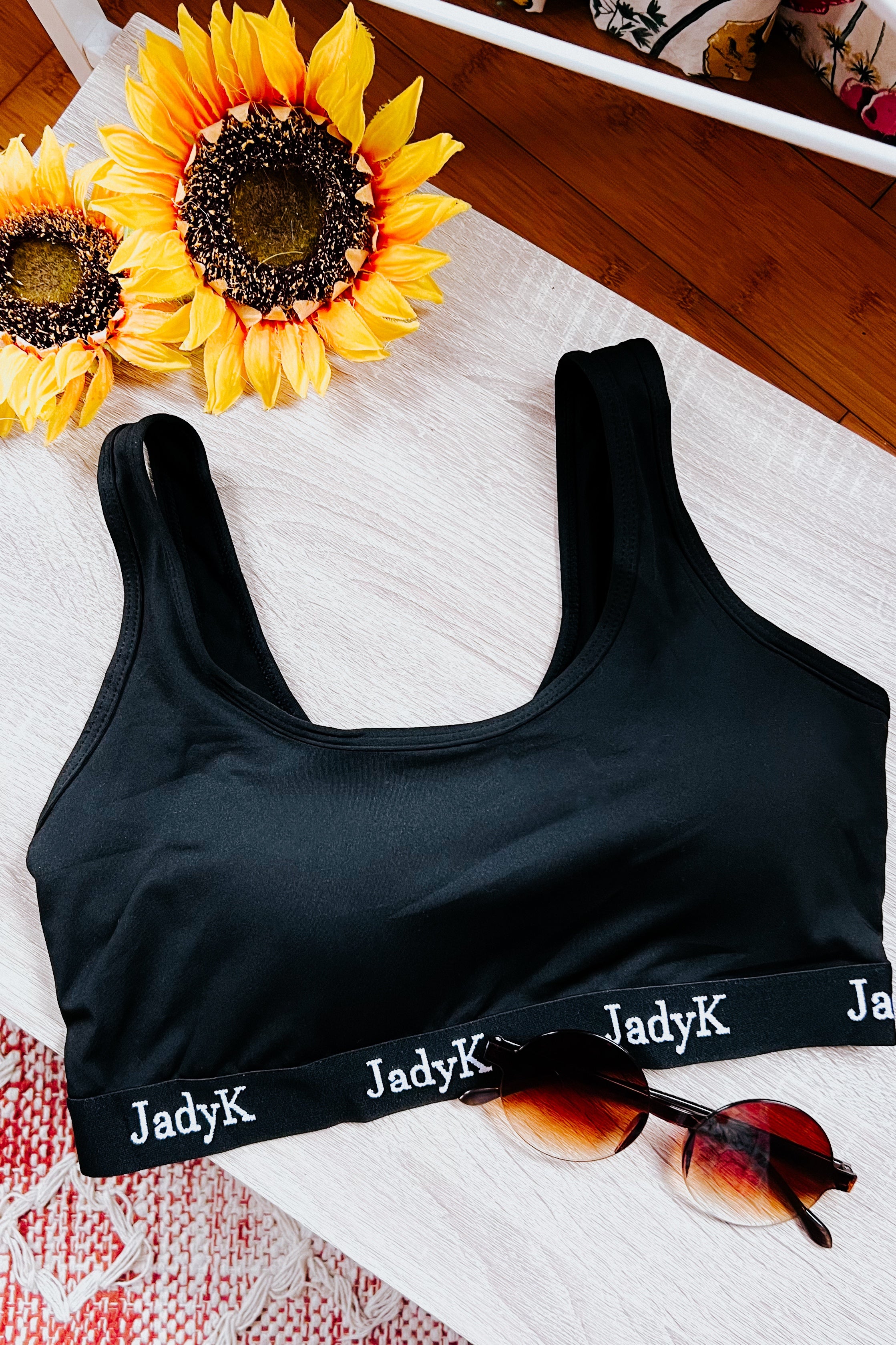 Black scoop bralette with JadyK logo band styled with tortoiseshell sunglasses and artificial sunflowers on wooden surface