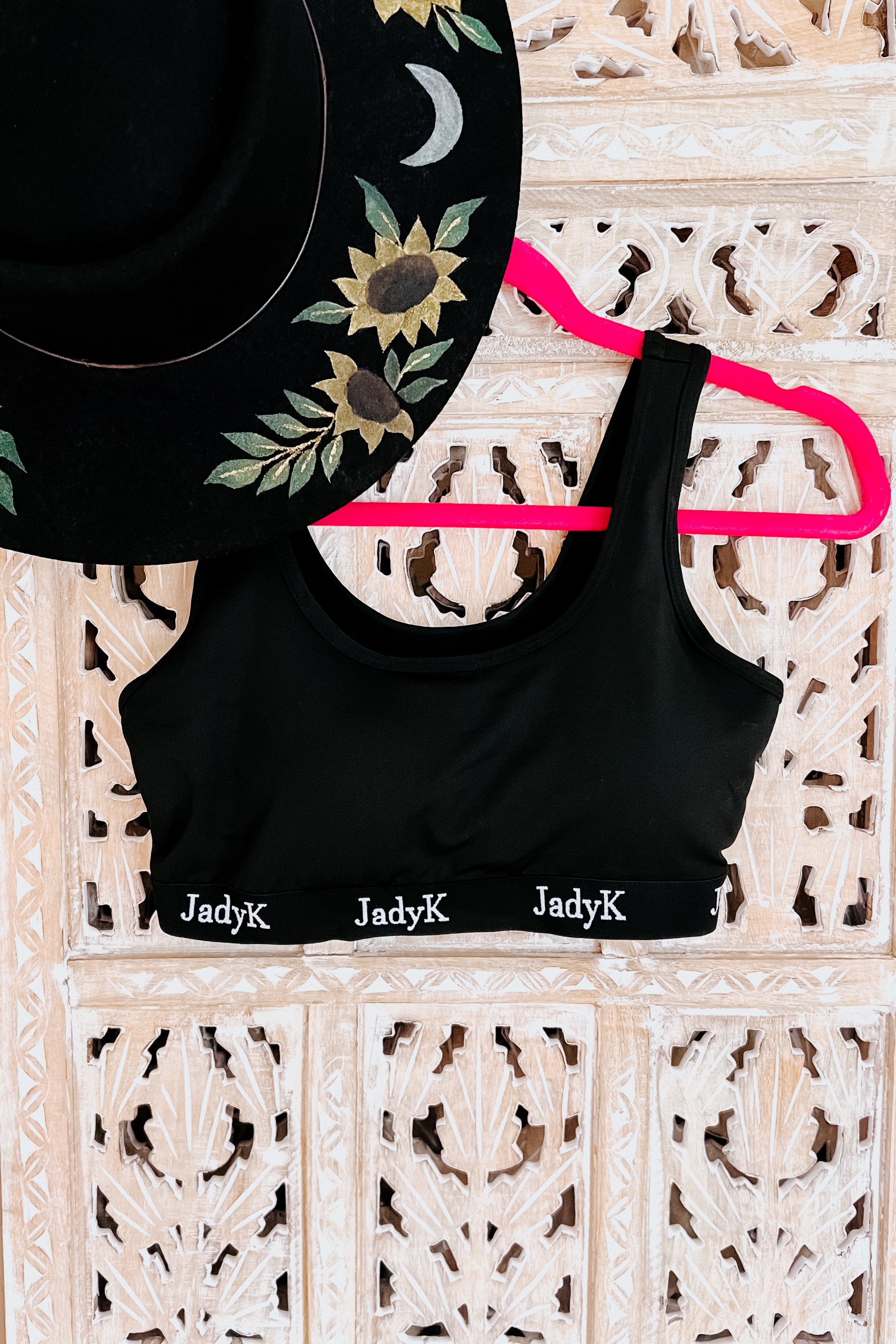 Black scoop bralette with branded elastic band hanging on bright pink hanger next to black embroidered sunflower hat