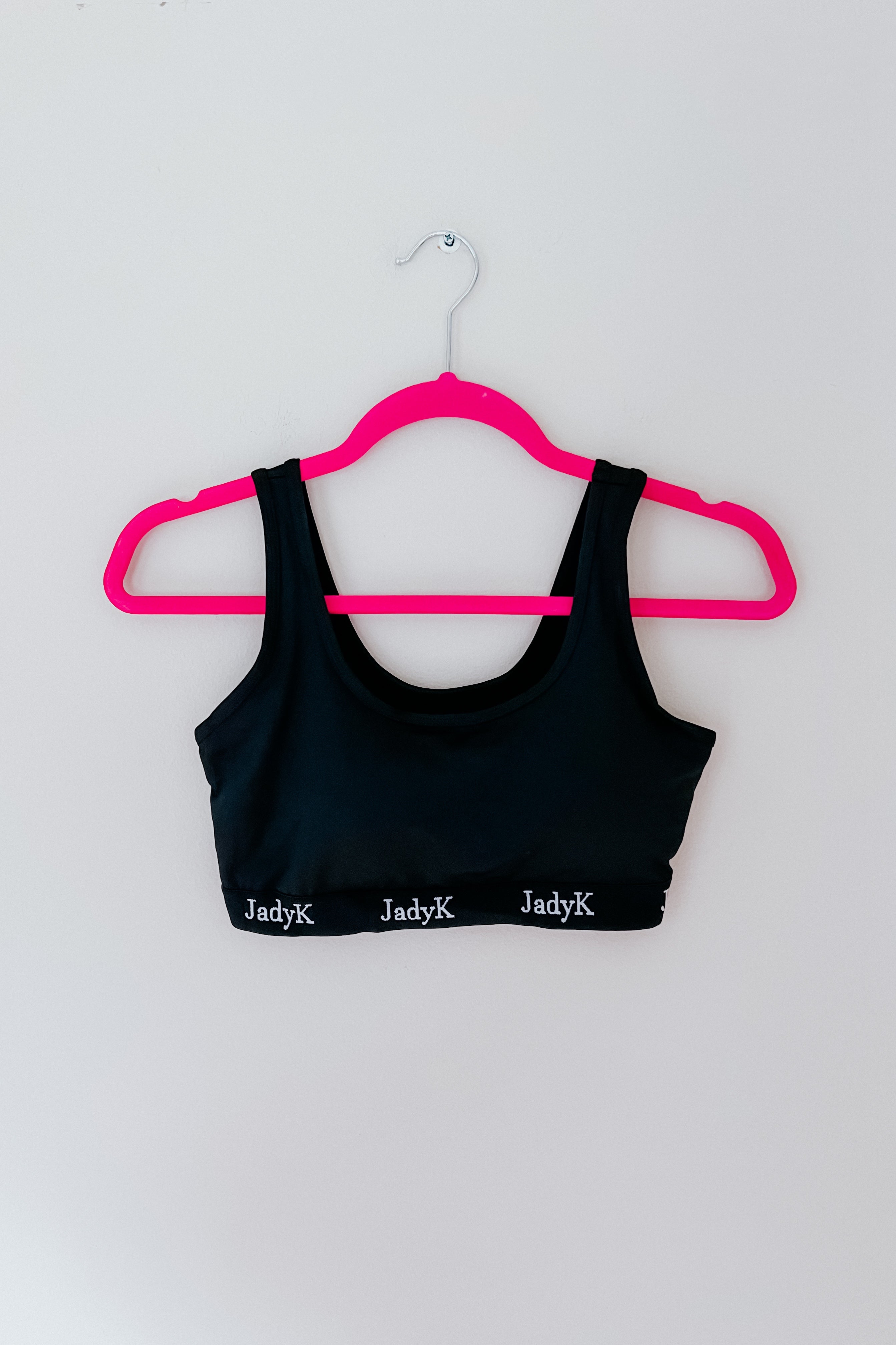 Black scoop bralette with repeated white logo on band hanging on bright pink hanger against plain white wall