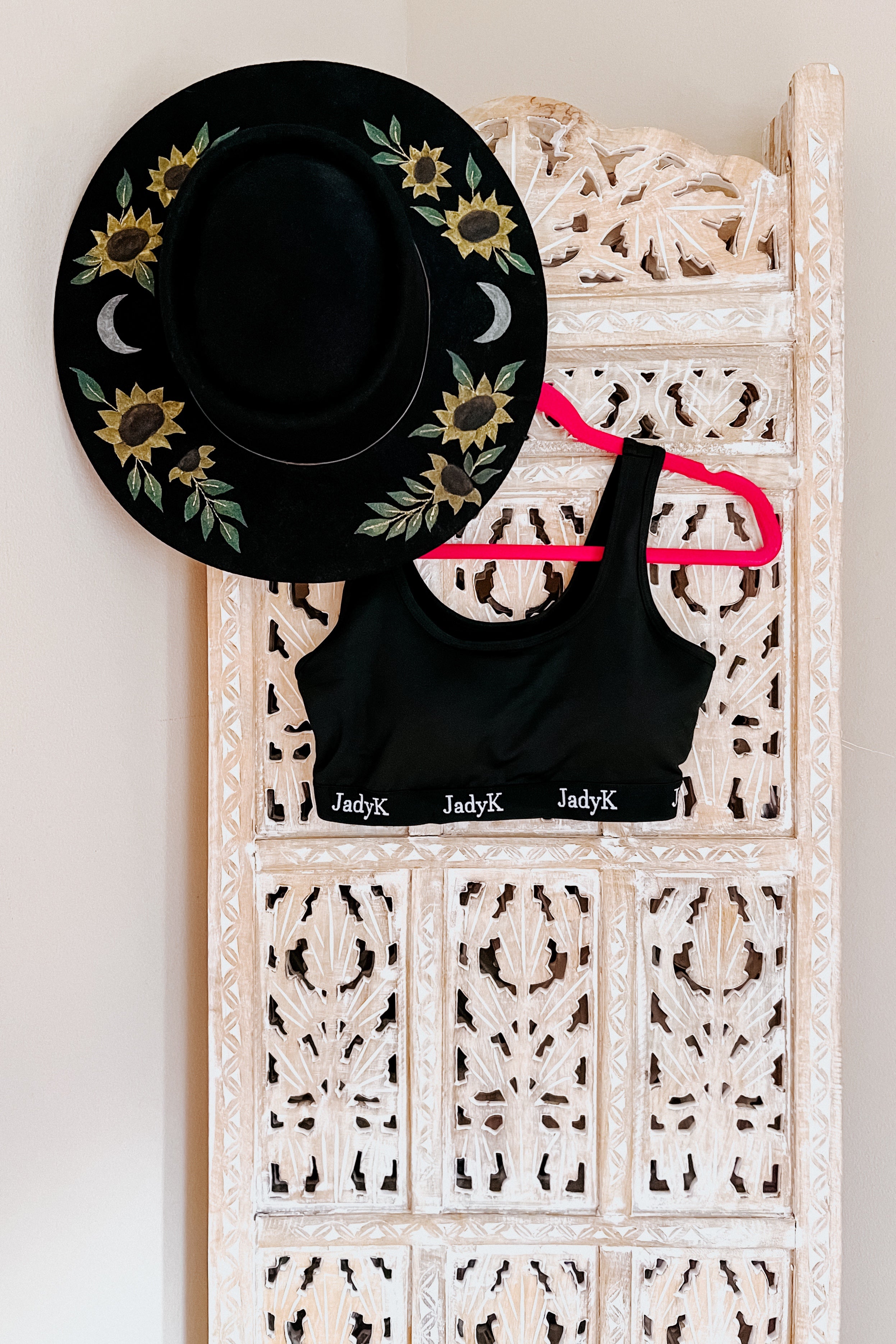 Black scoop bralette with white logo on band hanging on bright pink hanger beside black hat with yellow sunflower and crescent moon embroidery