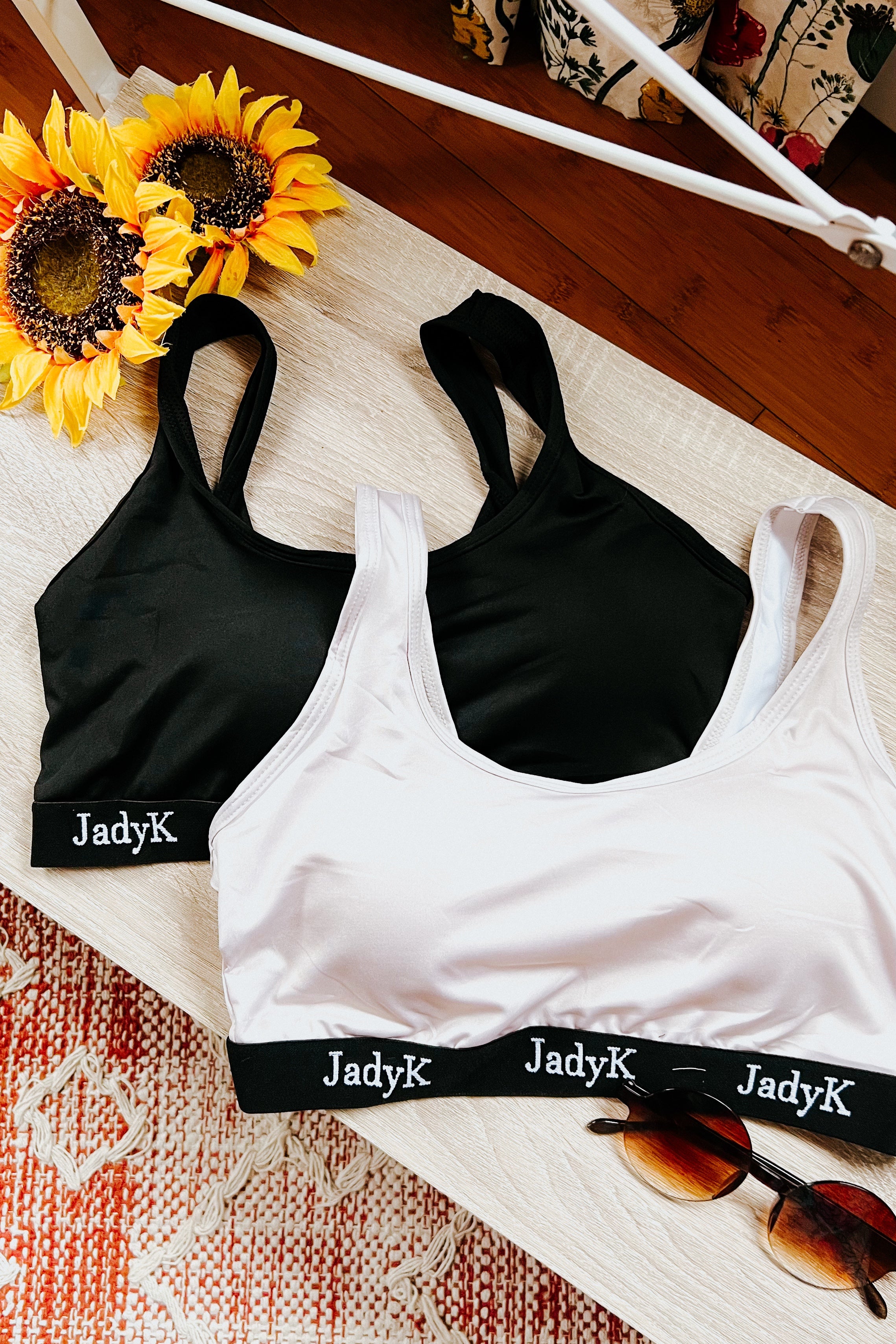 Two scoop bralettes in black and champagne with branded elastic bands displayed beside sunflowers and sunglasses