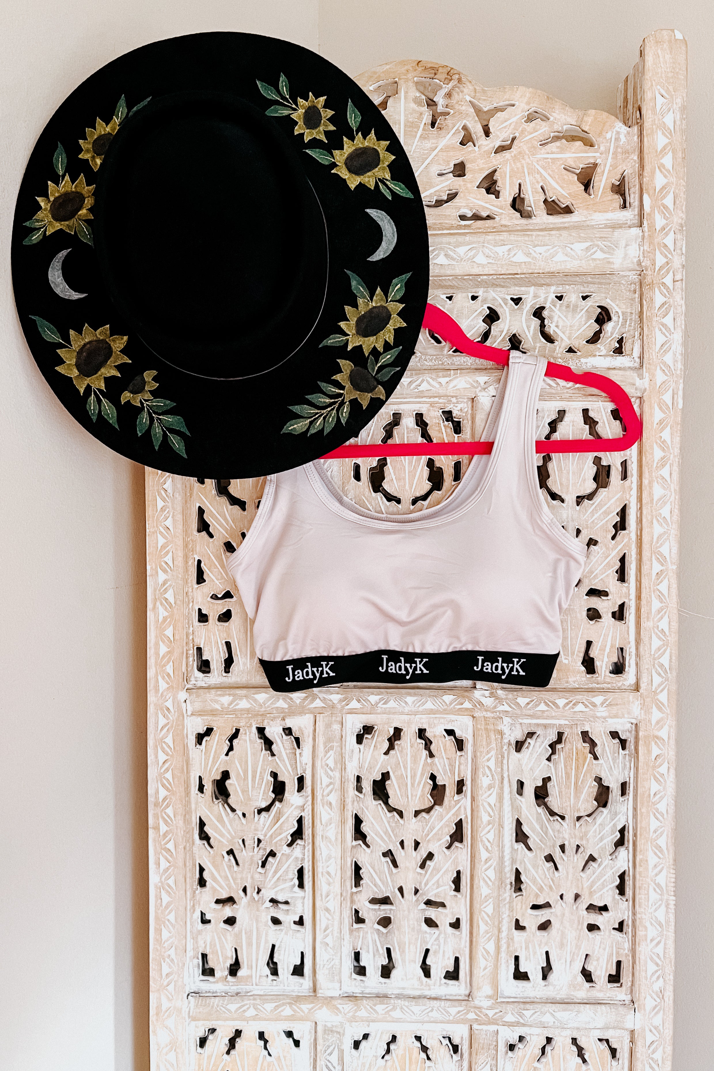Champagne scoop bralette with black branded band hanging on carved wooden screen beside black hat with sunflower embroidery