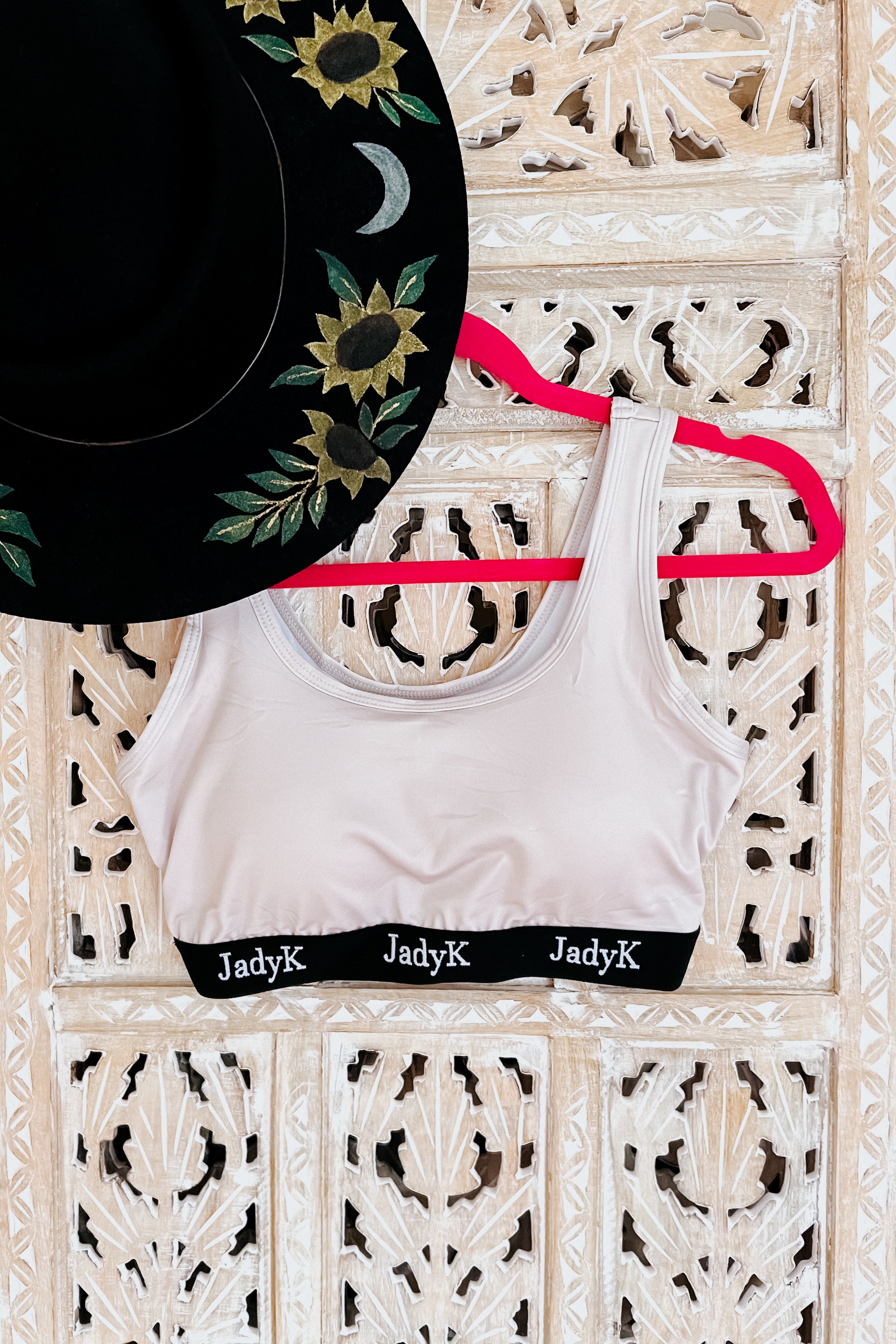 Champagne bralette with black branded band hanging on pink hanger beside black hat with sunflower and crescent moon embroidery