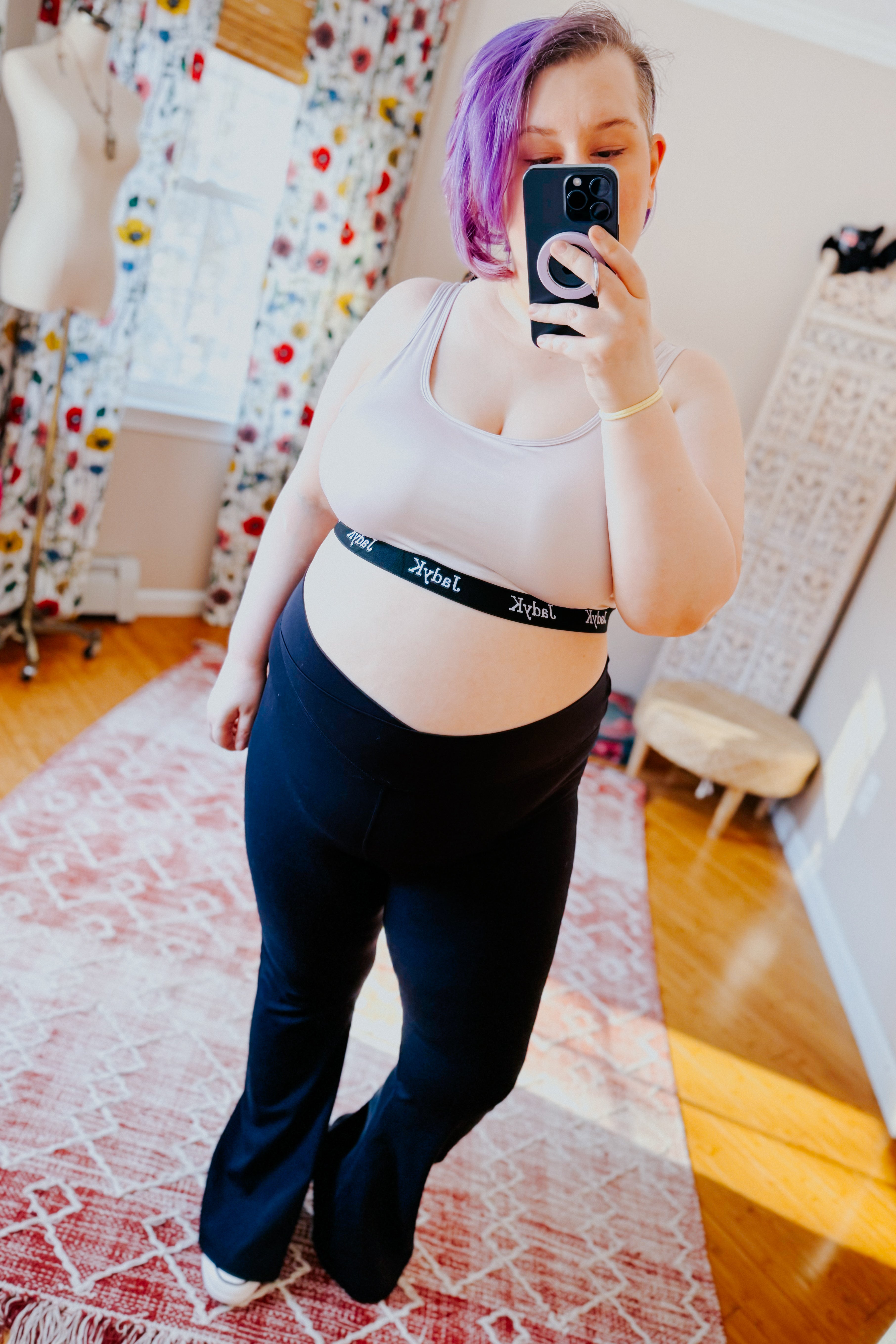 Woman with purple hair wearing champagne bralette with black branded band and black pants, taking a mirror selfie indoors