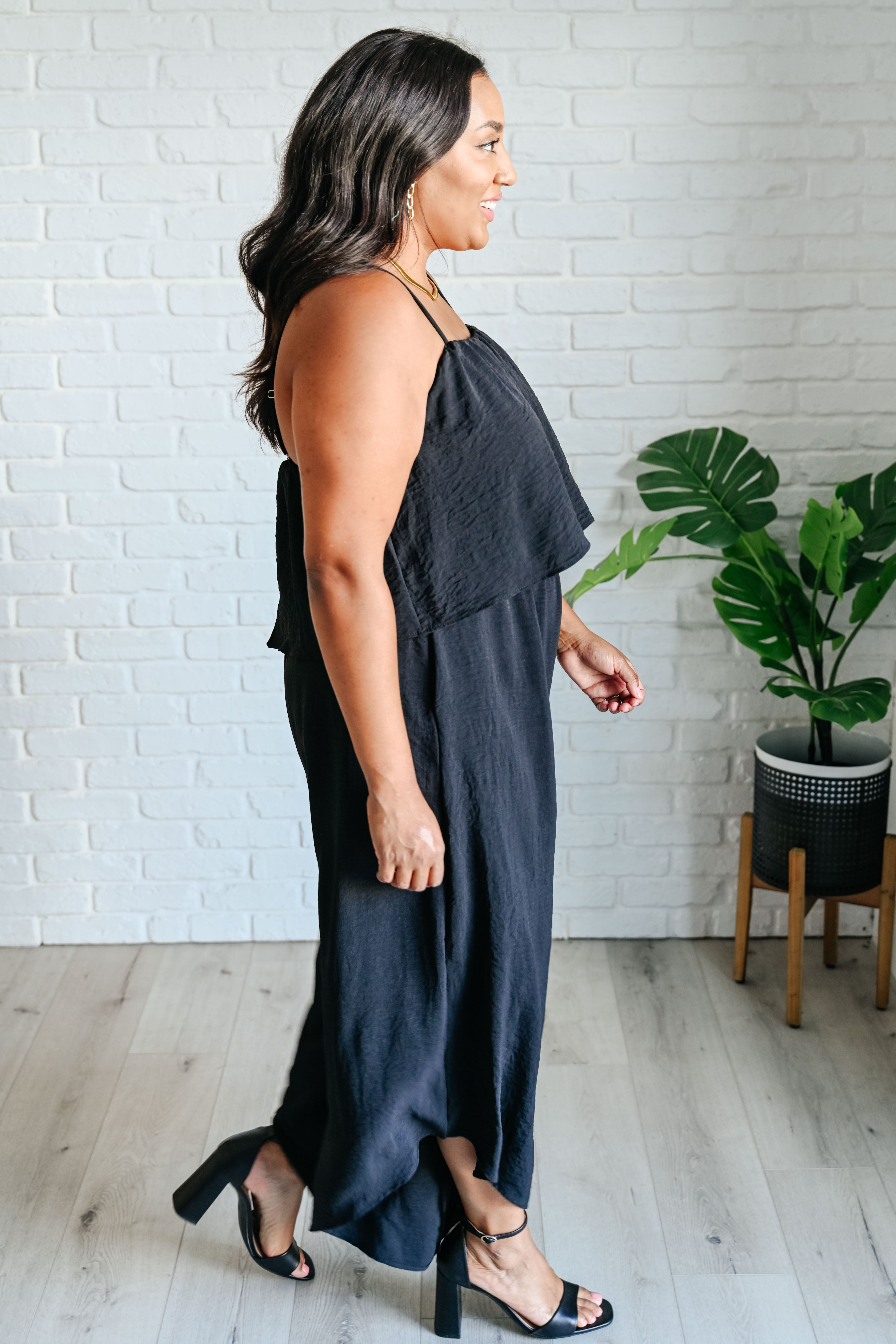 Profile view of a woman wearing a black sleeveless jumpsuit with thin straps and block heel sandals indoors