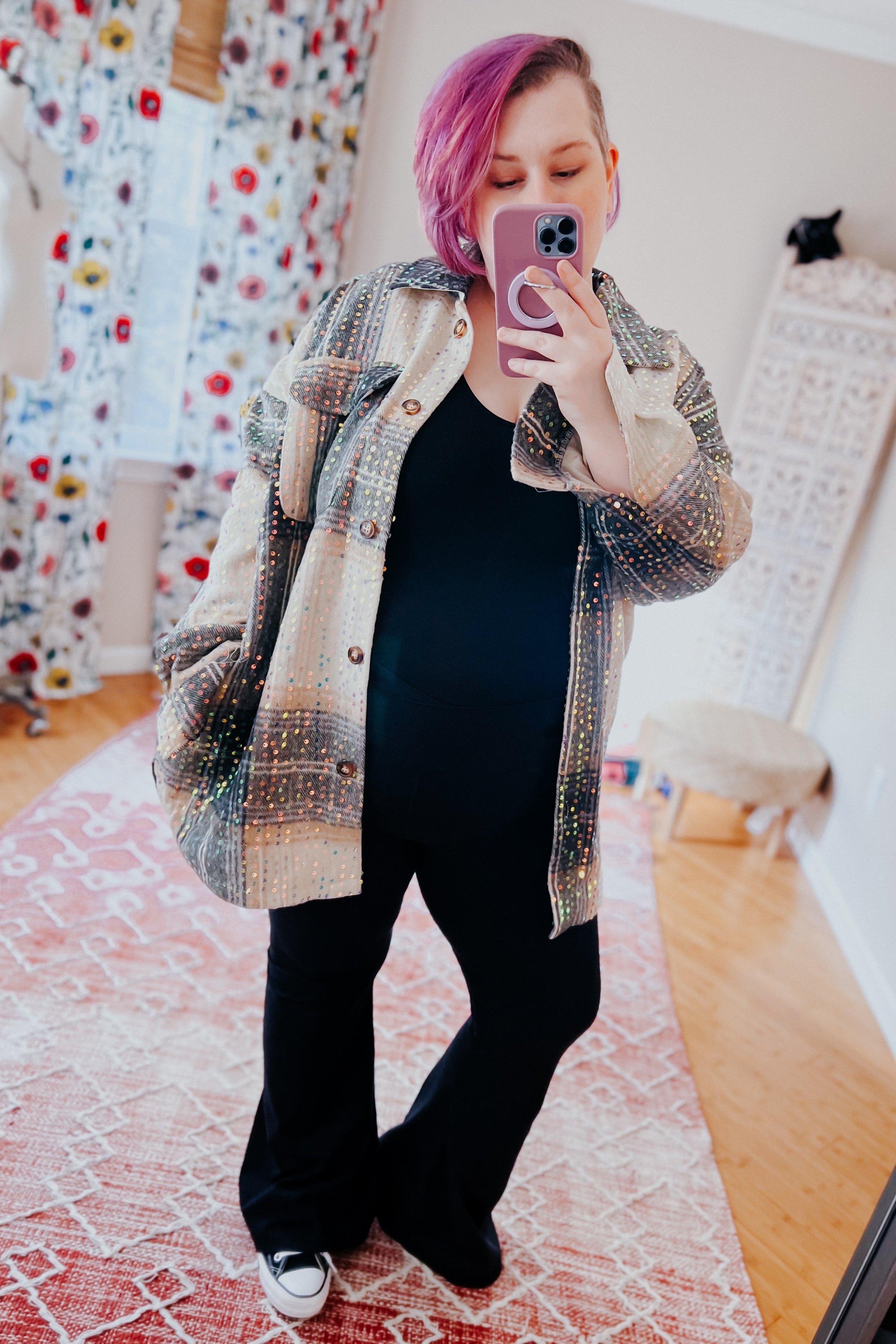 Woman with purple hair wearing sequined beige and black plaid shacket over black outfit taking a mirror selfie indoors