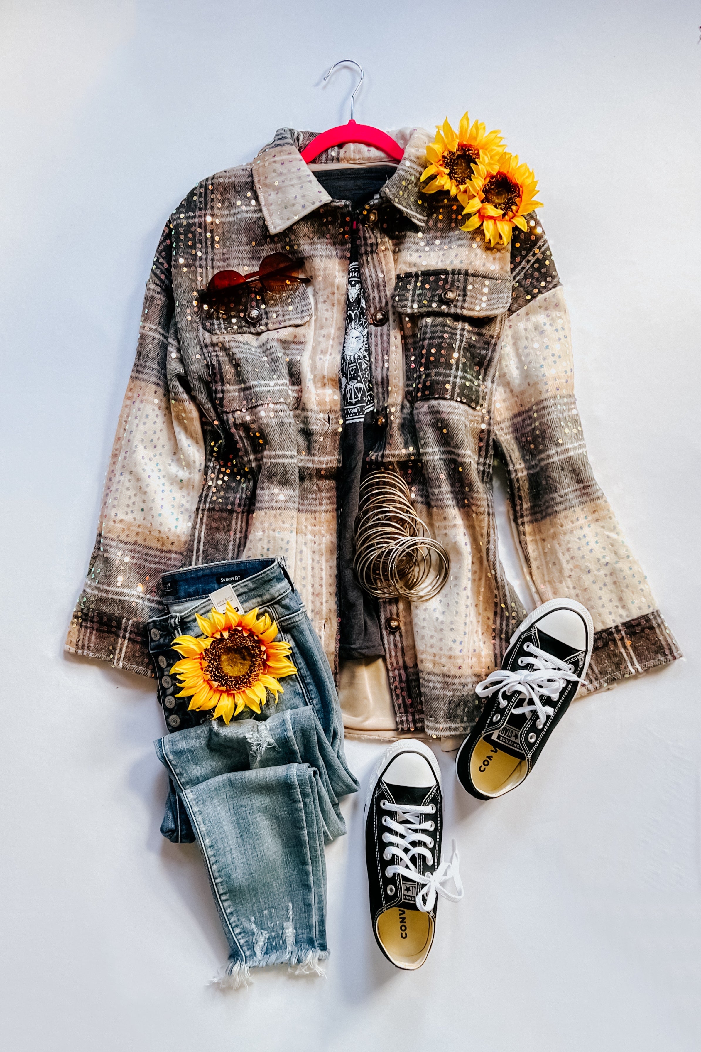 Beige and black sequin plaid shacket styled with faded jeans, black sneakers, sunflower accents, and layered bracelets on white background