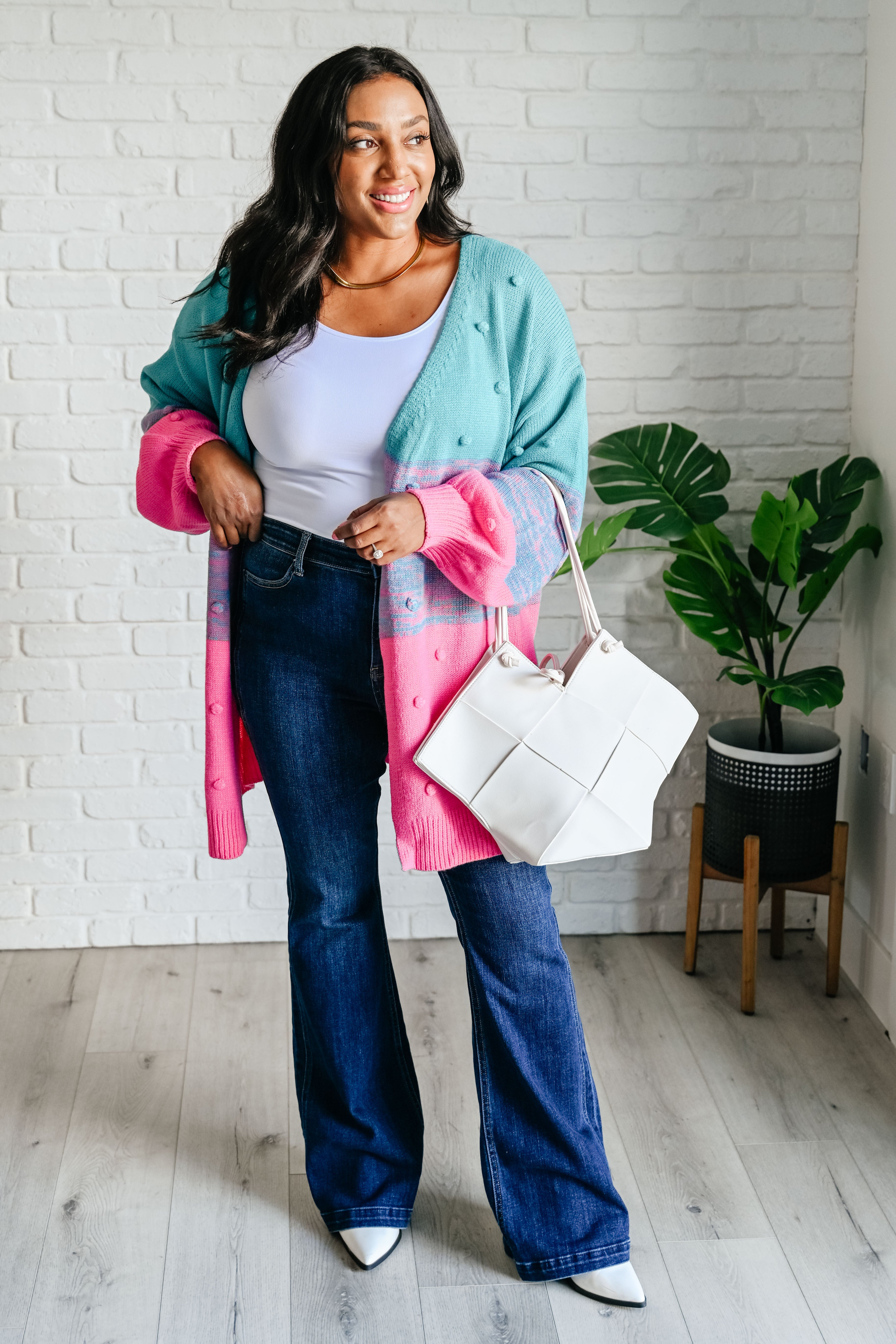 Woman in teal and pink open front cardigan paired with white top, dark jeans, white boots, and white woven tote bag
