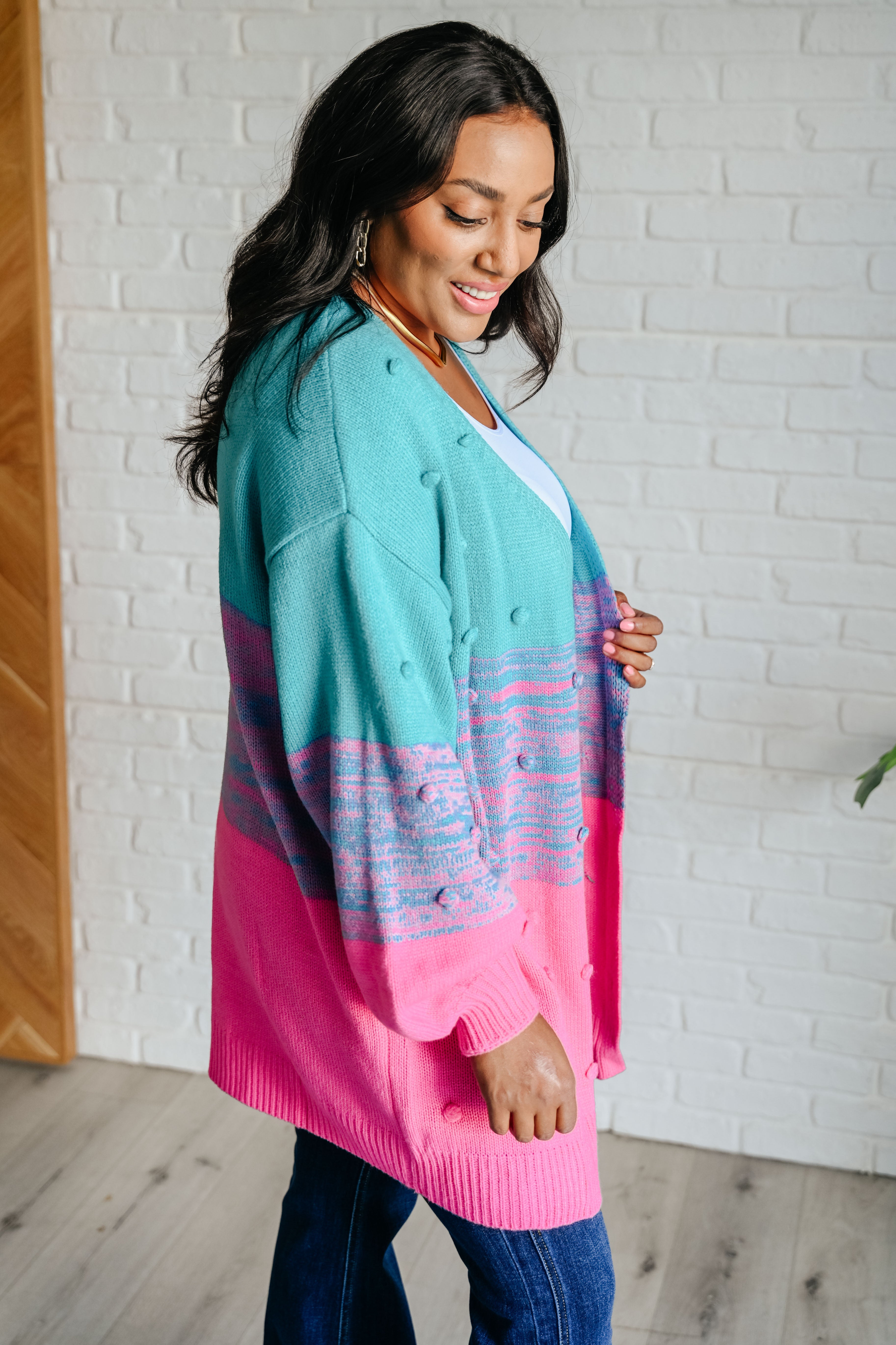 Woman wearing teal and pink open front cardigan with textured bobbles, paired with white top and blue jeans in bright room