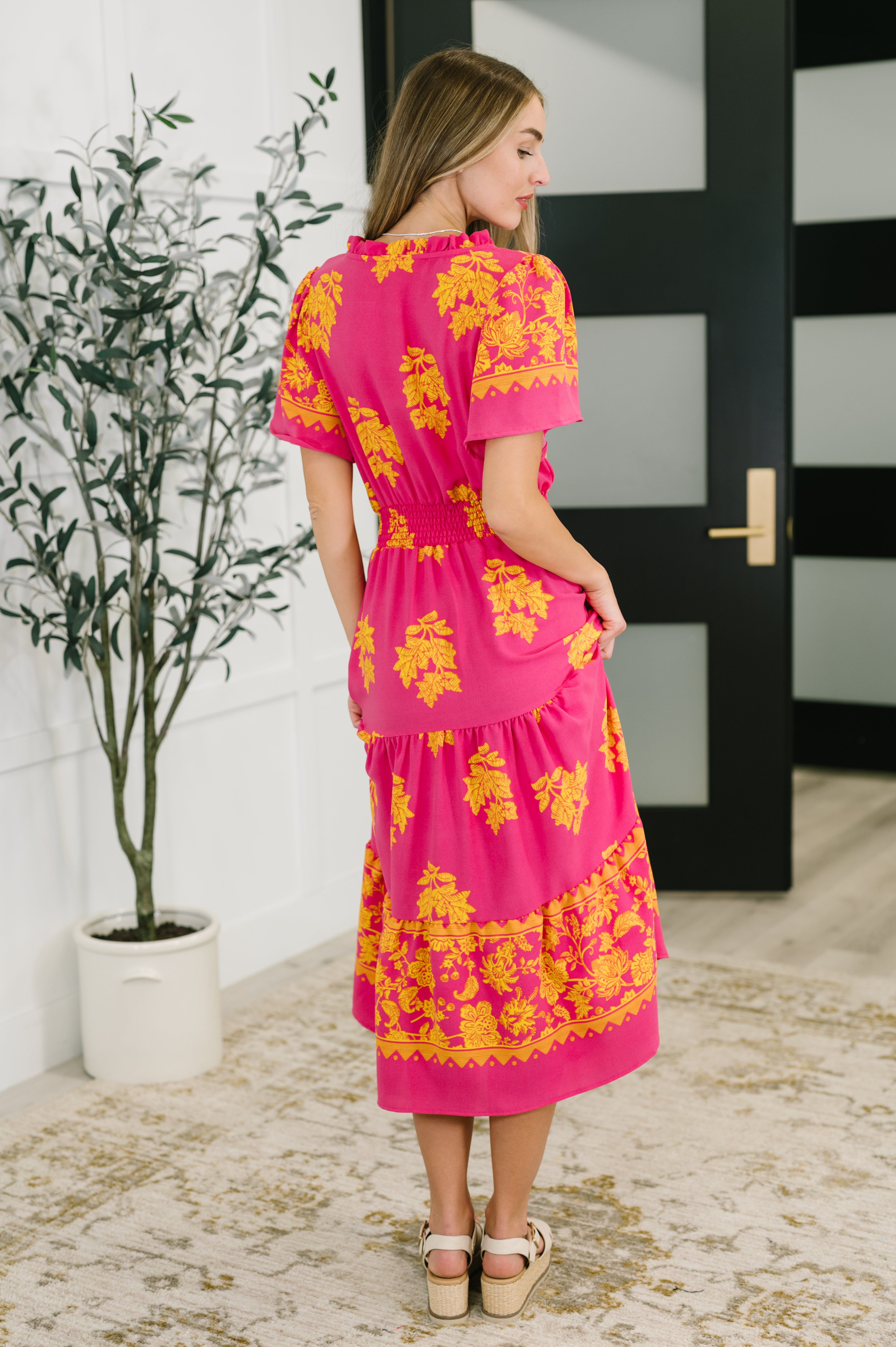 Woman indoors wearing fuchsia V-neck dress with yellow floral print and elastic waist standing with hands in pockets