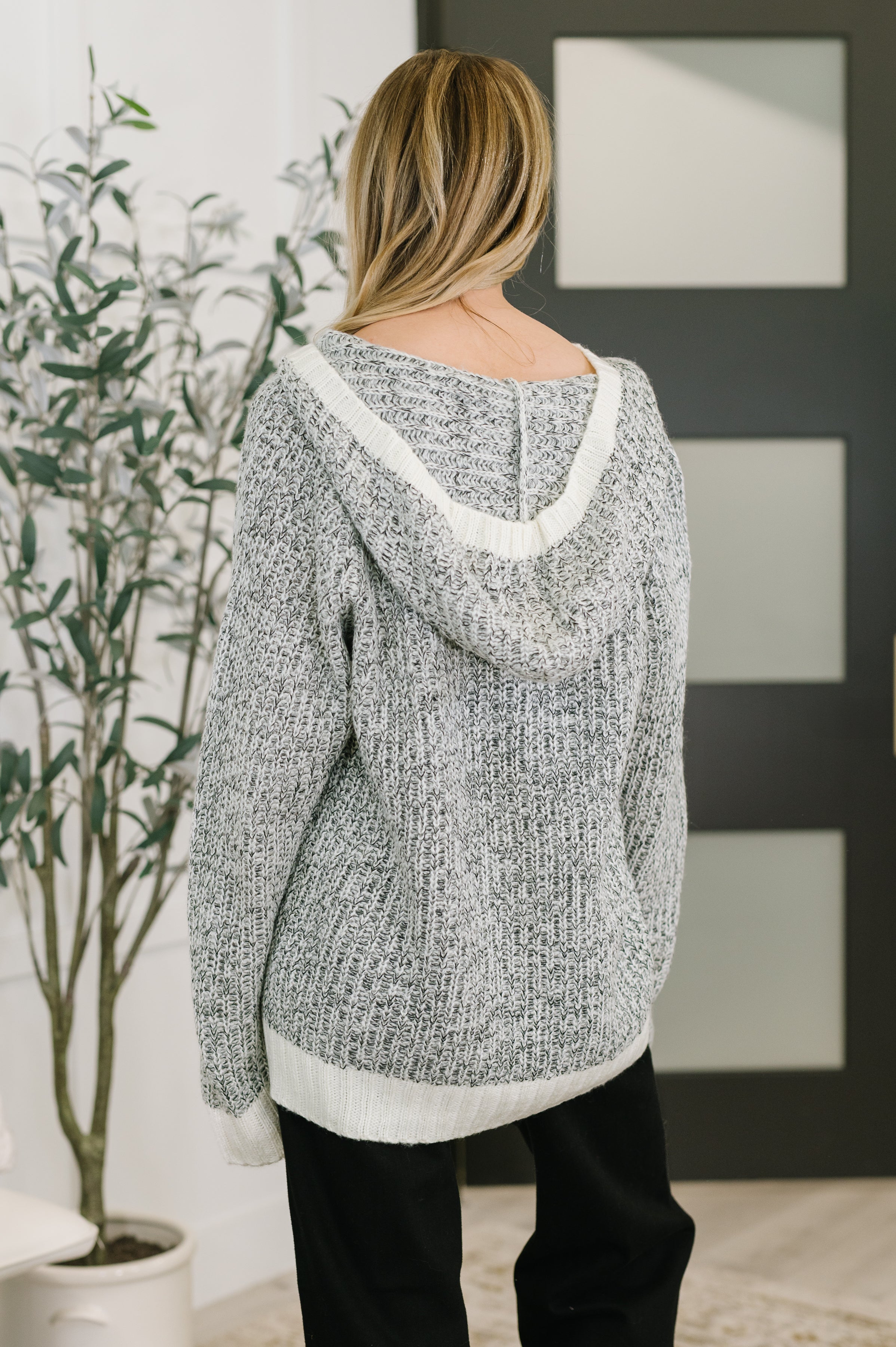 Woman wearing grey textured knit pullover with hood and white trim, standing indoors near plant and modern door