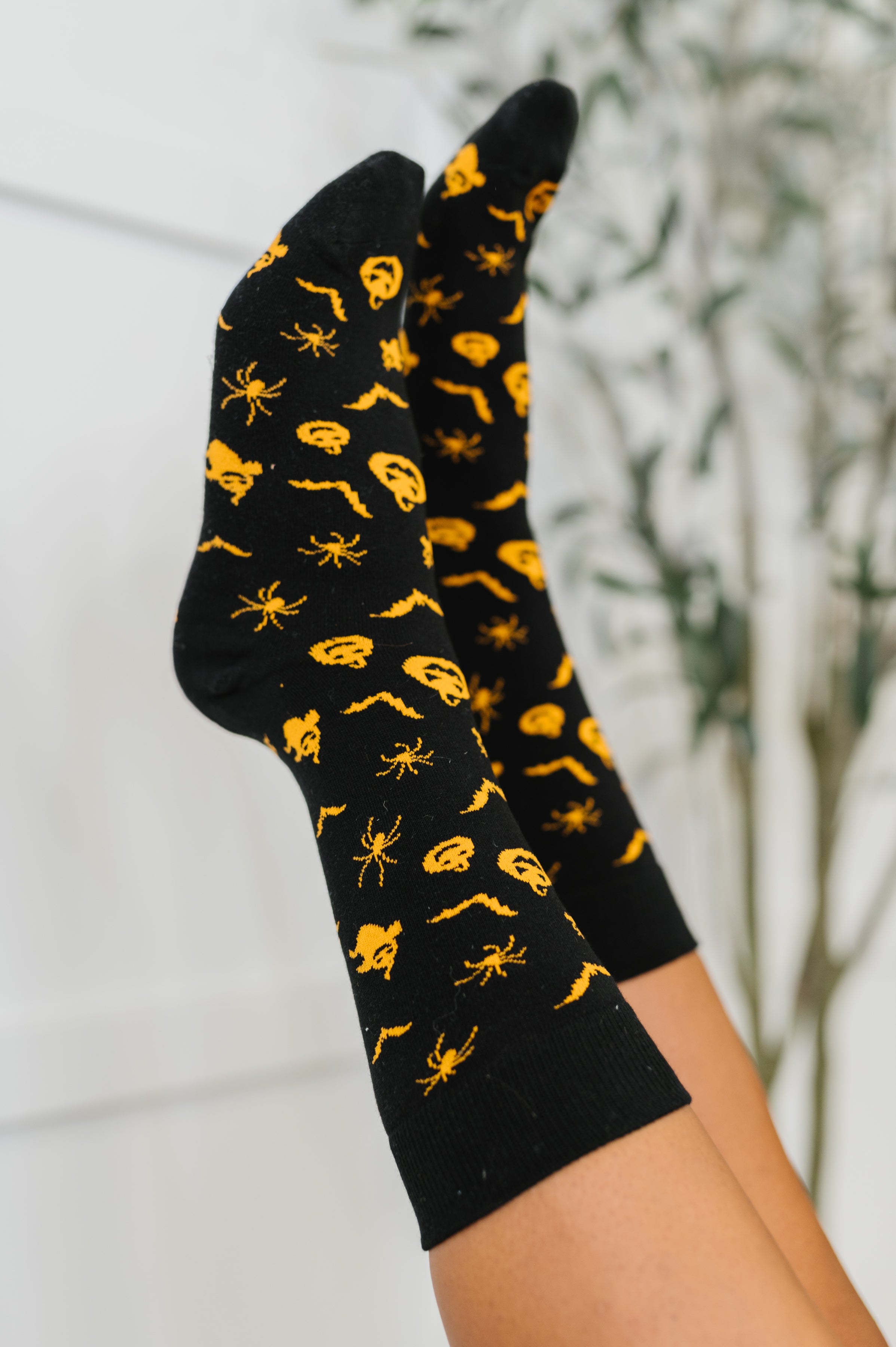 Black quarter socks featuring bright orange spooky designs worn indoors with blurred plant background