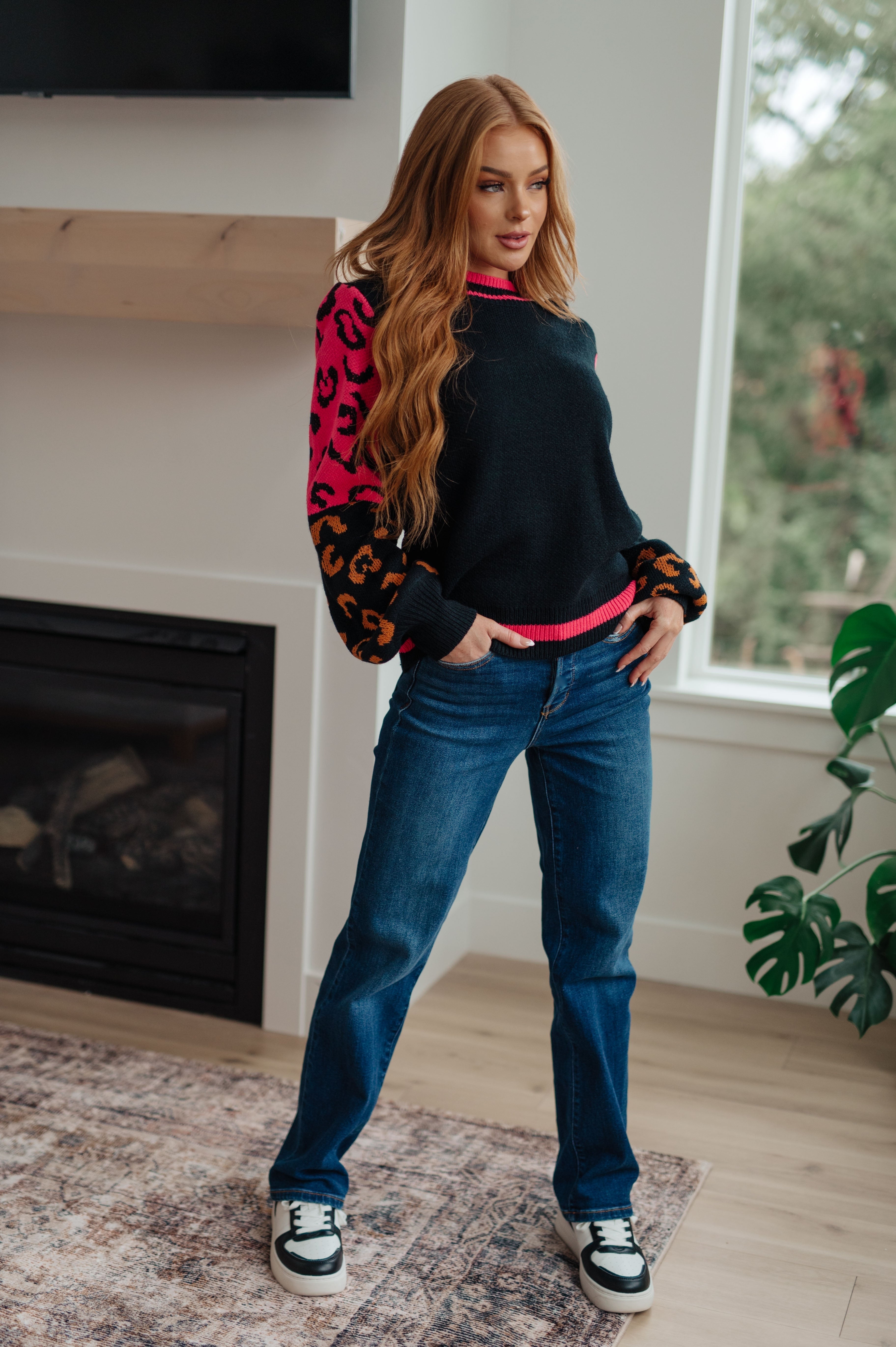 Woman wearing black sweater with bold pink and orange animal print sleeves paired with blue jeans indoors