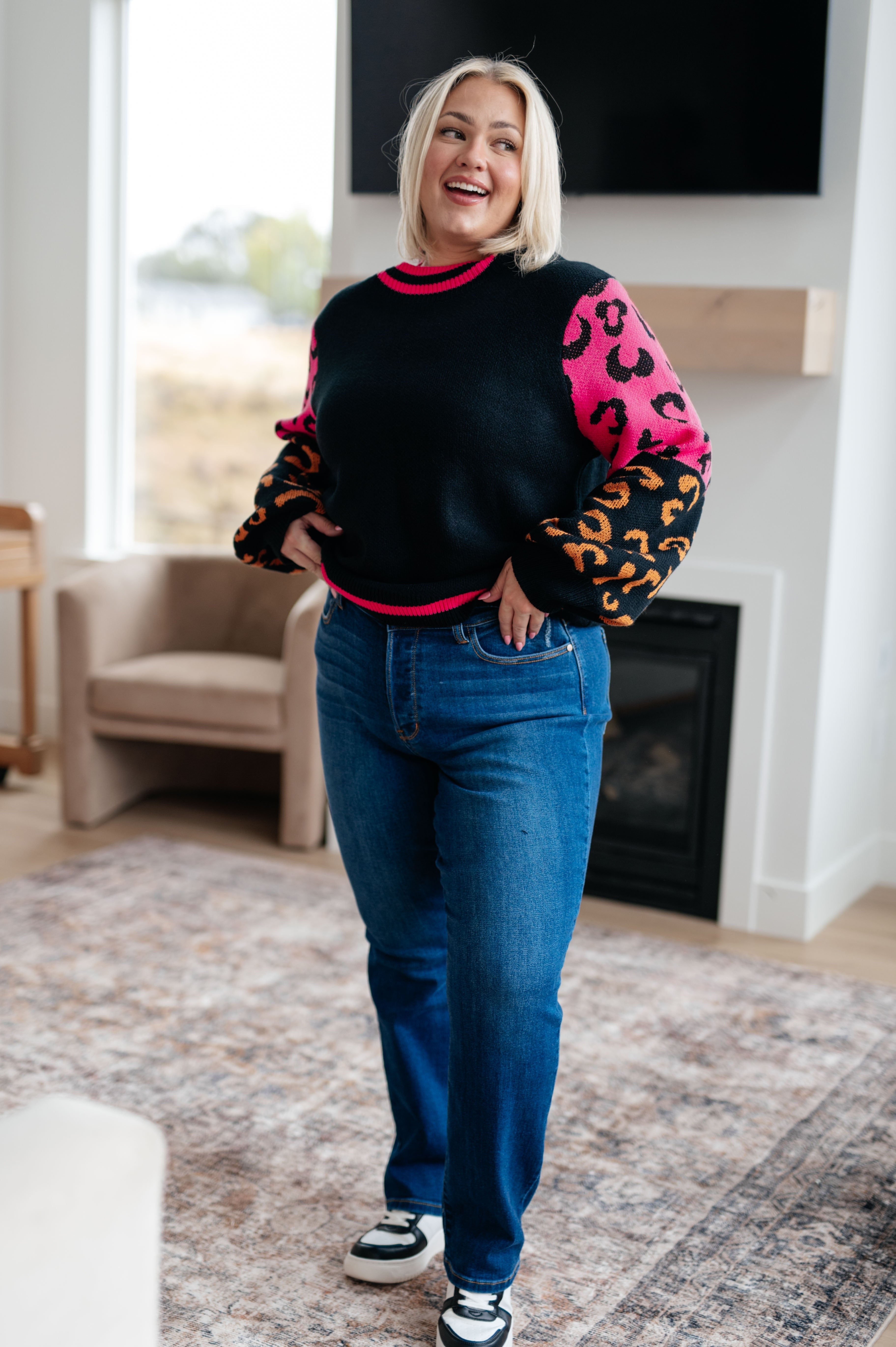 Woman wearing a black sweater with bold pink and orange animal print sleeves paired with blue jeans and sneakers indoors