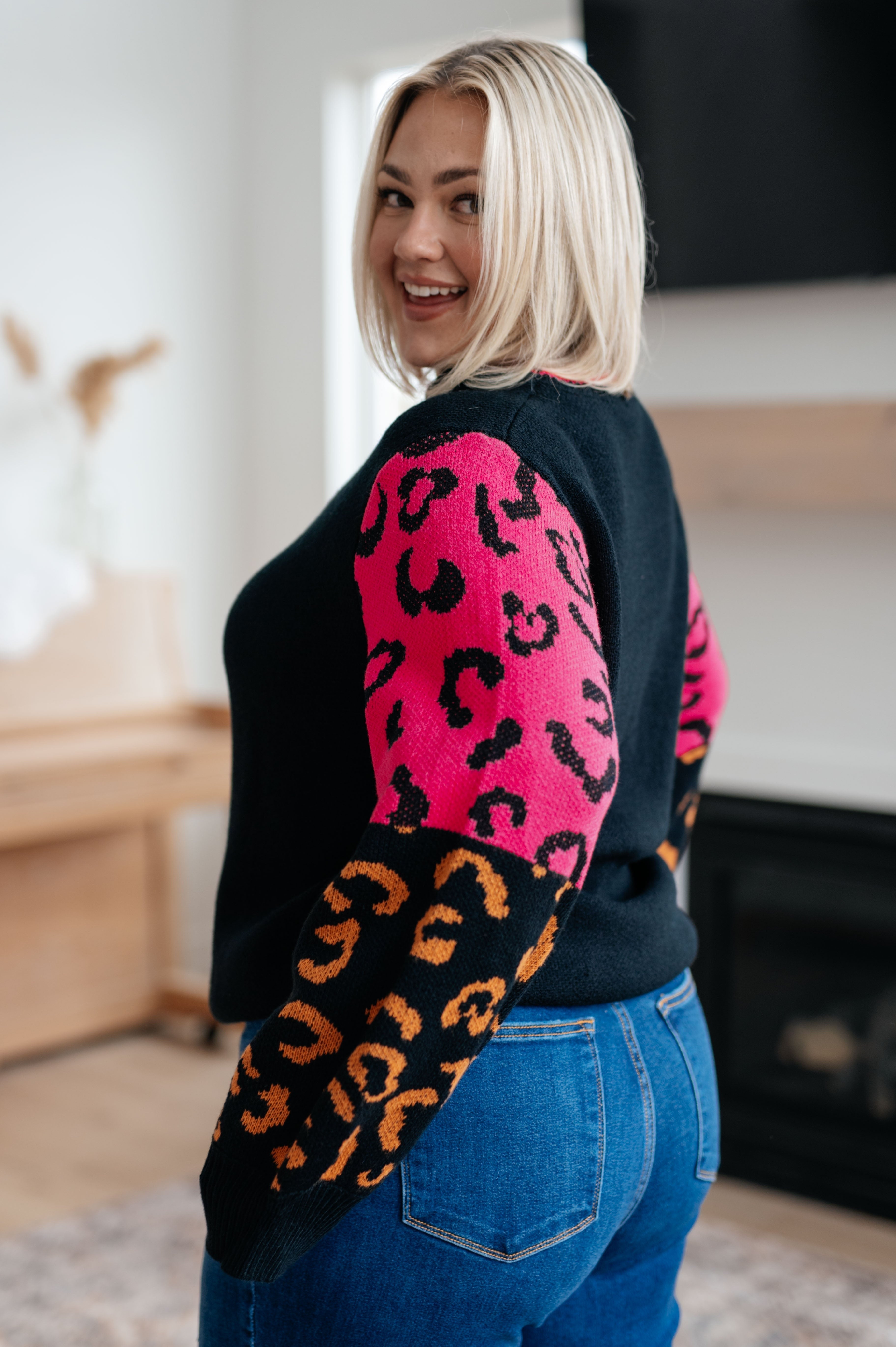 Woman smiling wearing black sweater with pink and orange animal print sleeves paired with blue jeans indoors