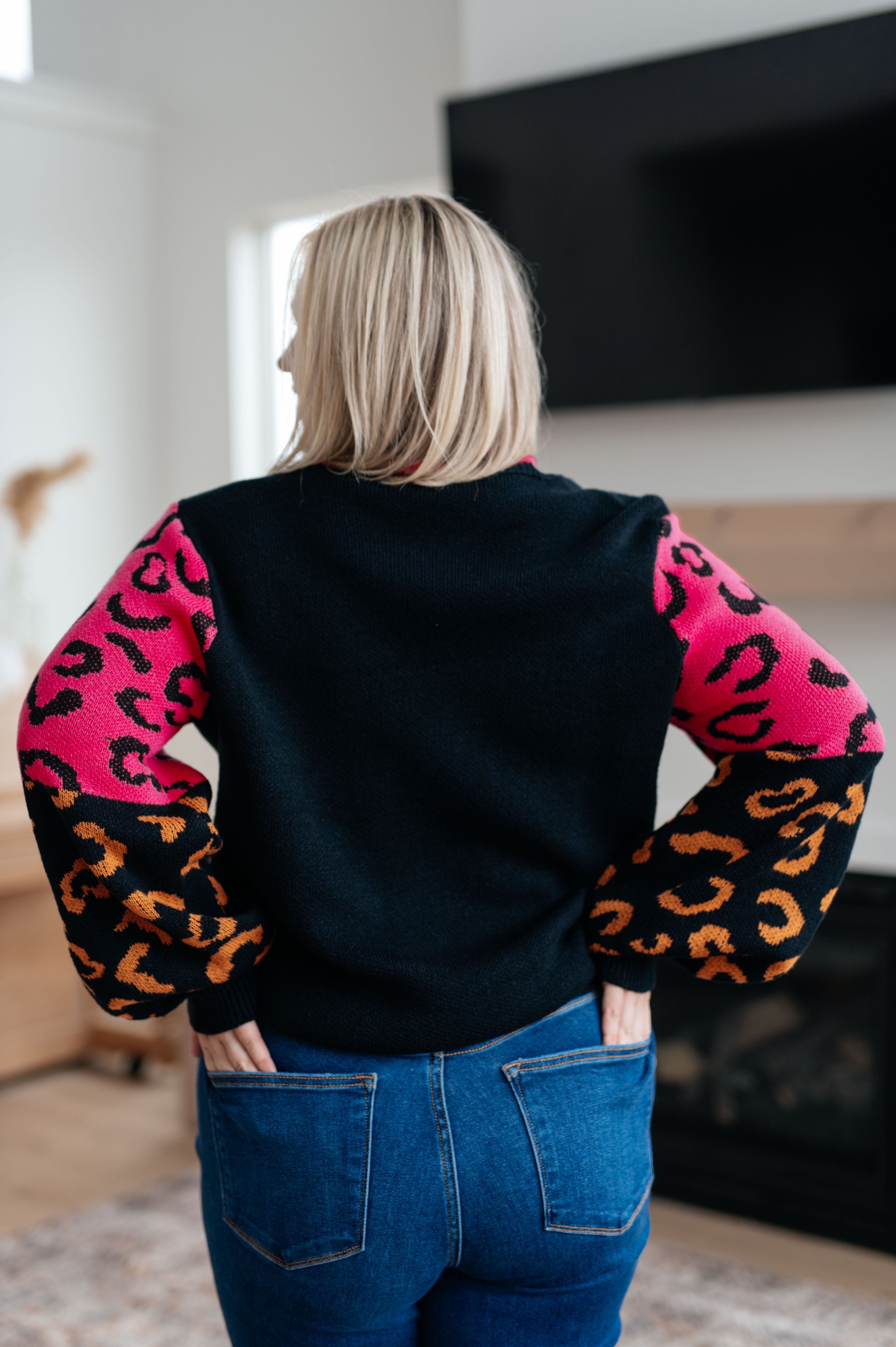 Woman wearing black sweater with bright pink and orange animal print sleeves, standing indoors facing away