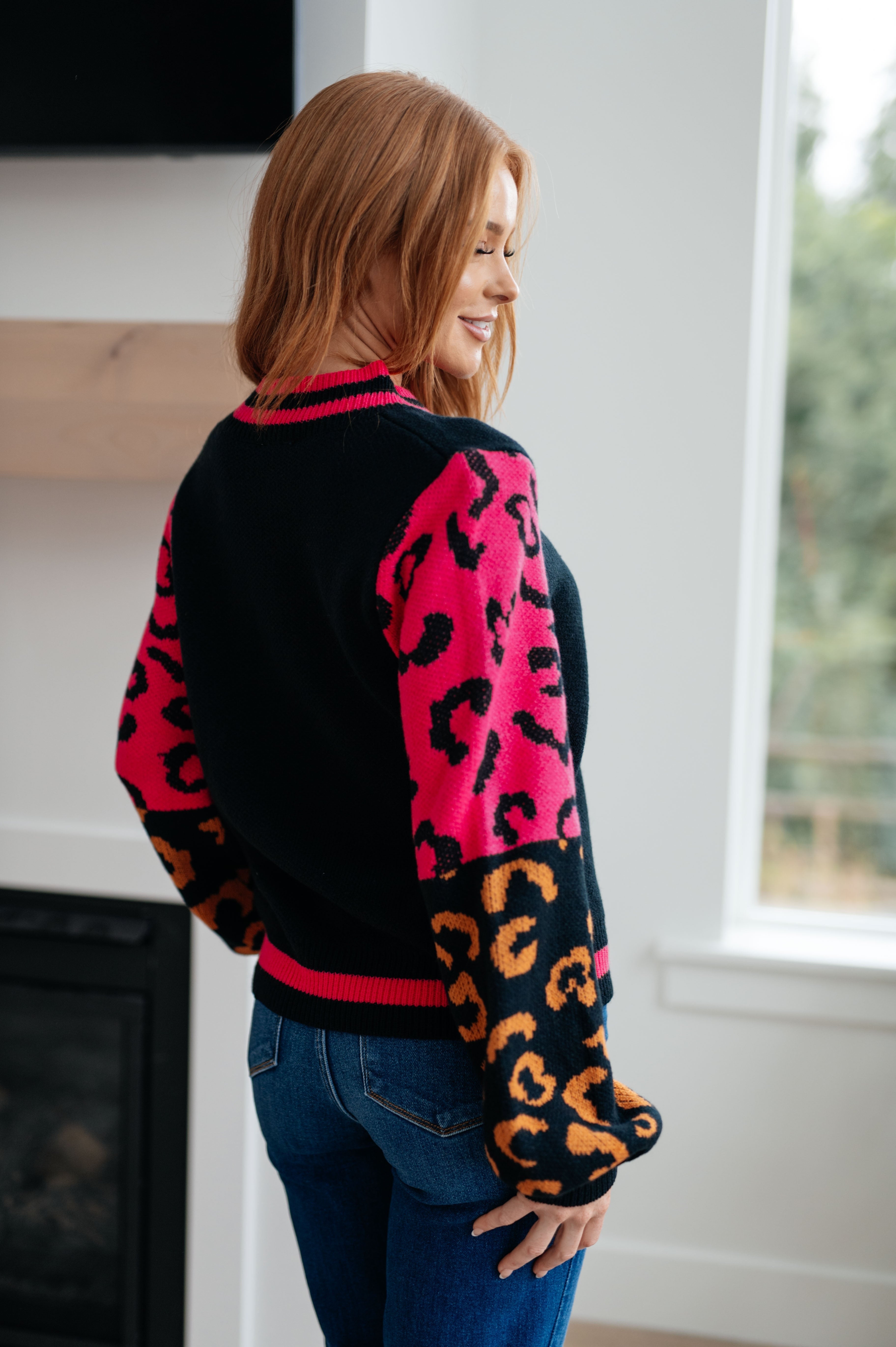 Woman wearing black sweater with bold pink and orange animal print sleeves standing near window indoors