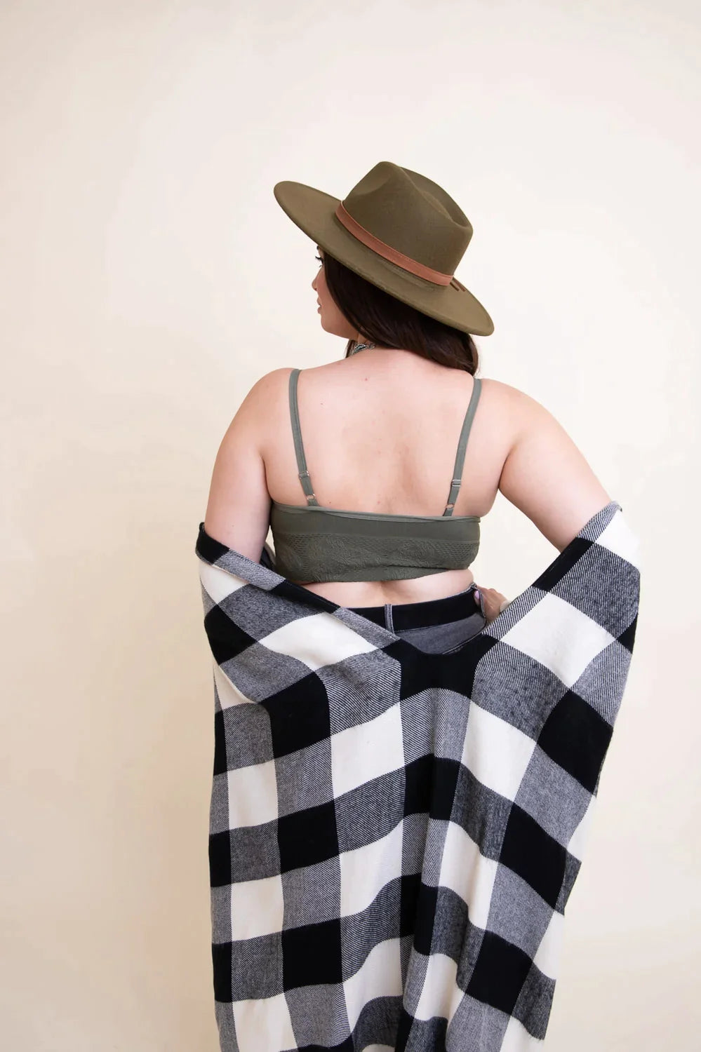 Woman wearing olive brami top and wide-brimmed hat, wrapped in black and white checkered blanket indoors