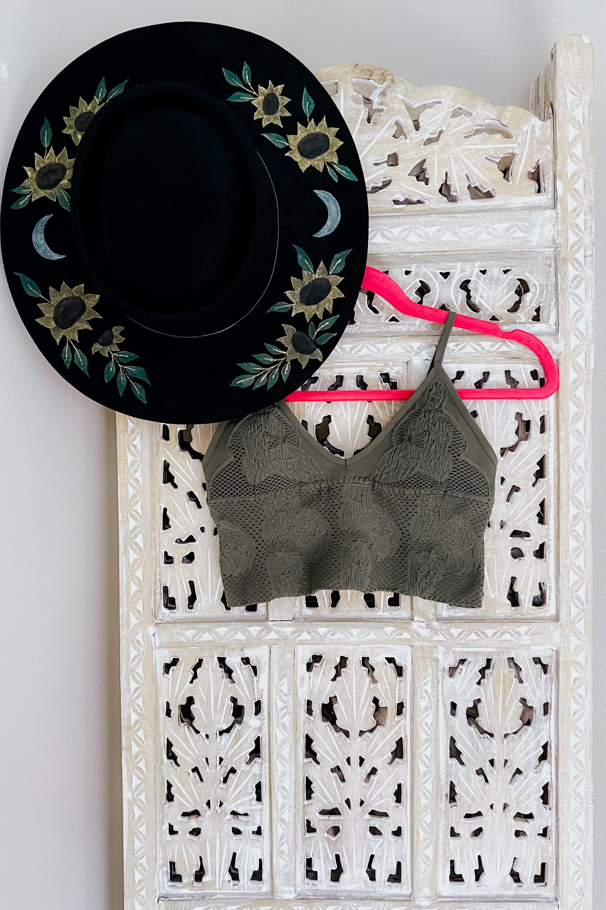 Olive textured brami with padding hanging on bright pink hanger beside black hat with floral and moon embroidery on carved screen