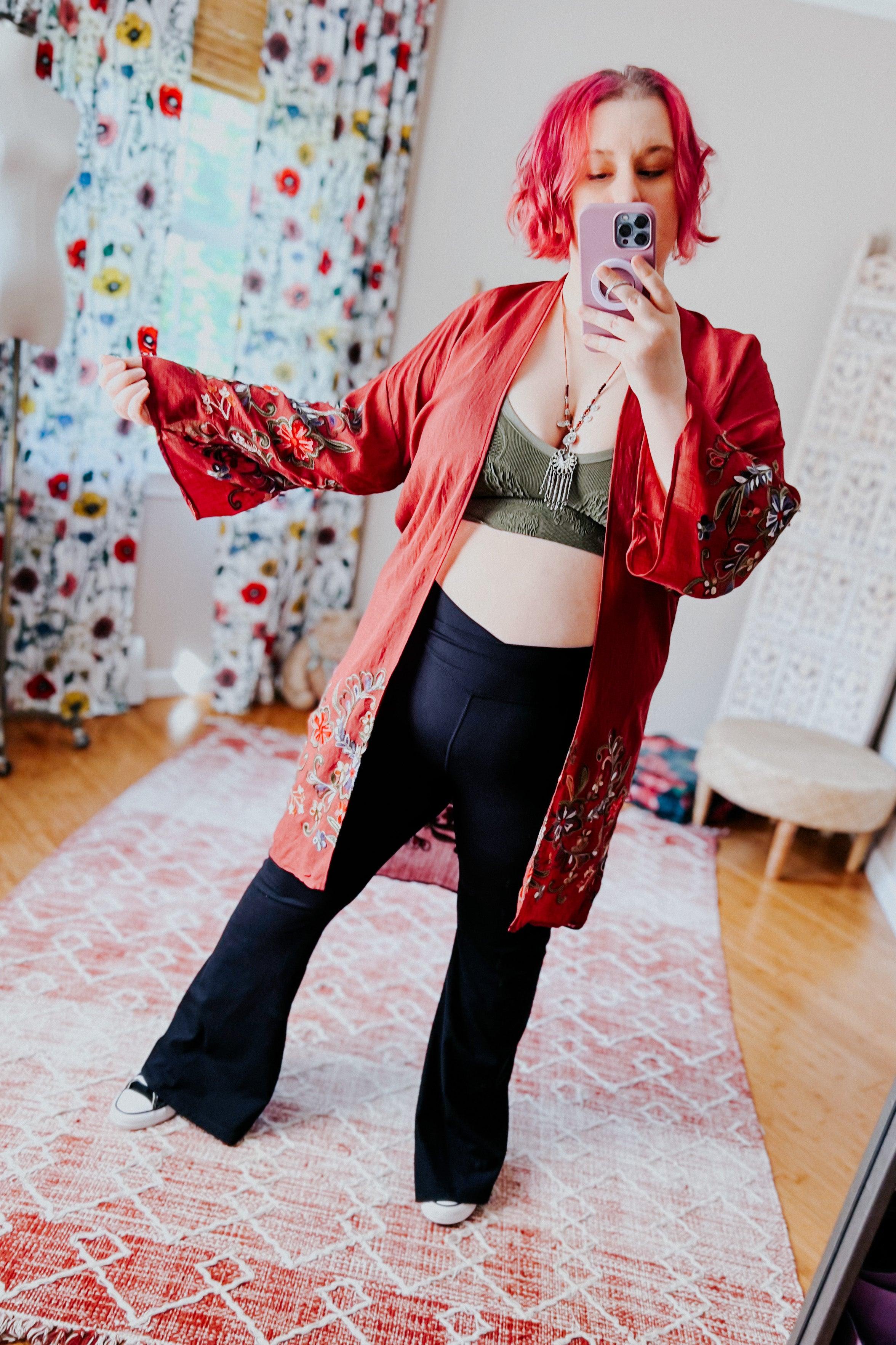 Woman with short pink hair wearing olive seamless padded brami and black pants, posing indoors with floral decor background