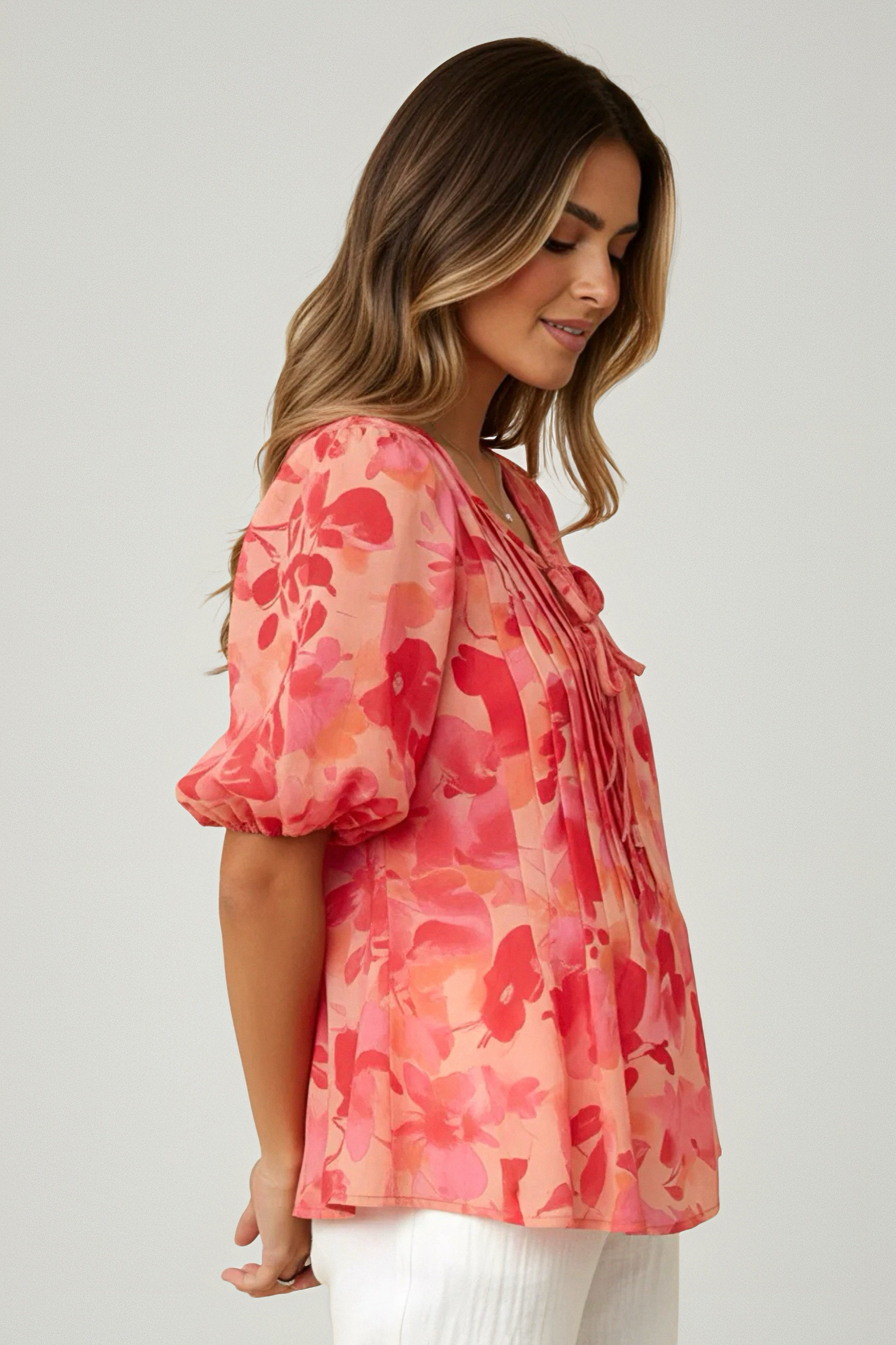 Woman wearing a peach blouse with red floral pattern and puff sleeves, paired with white pants, posing in profile view