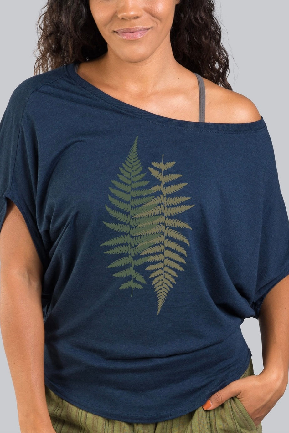 Woman wearing navy bamboo dolman top with fern graphic and off-shoulder neckline paired with striped olive pants