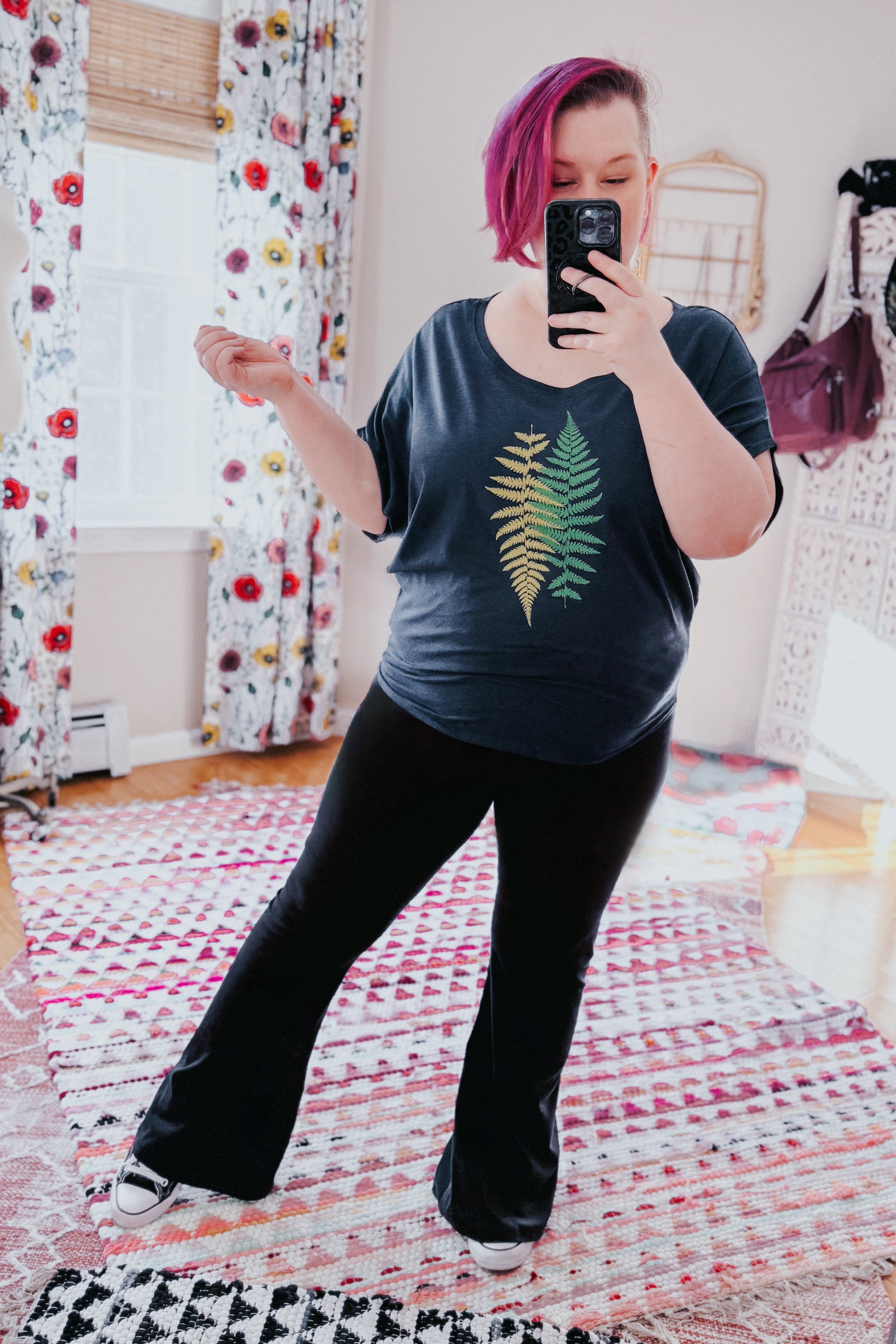 Woman with vibrant pink hair wearing navy top featuring fern design, black pants, and white shoes taking mirror selfie indoors