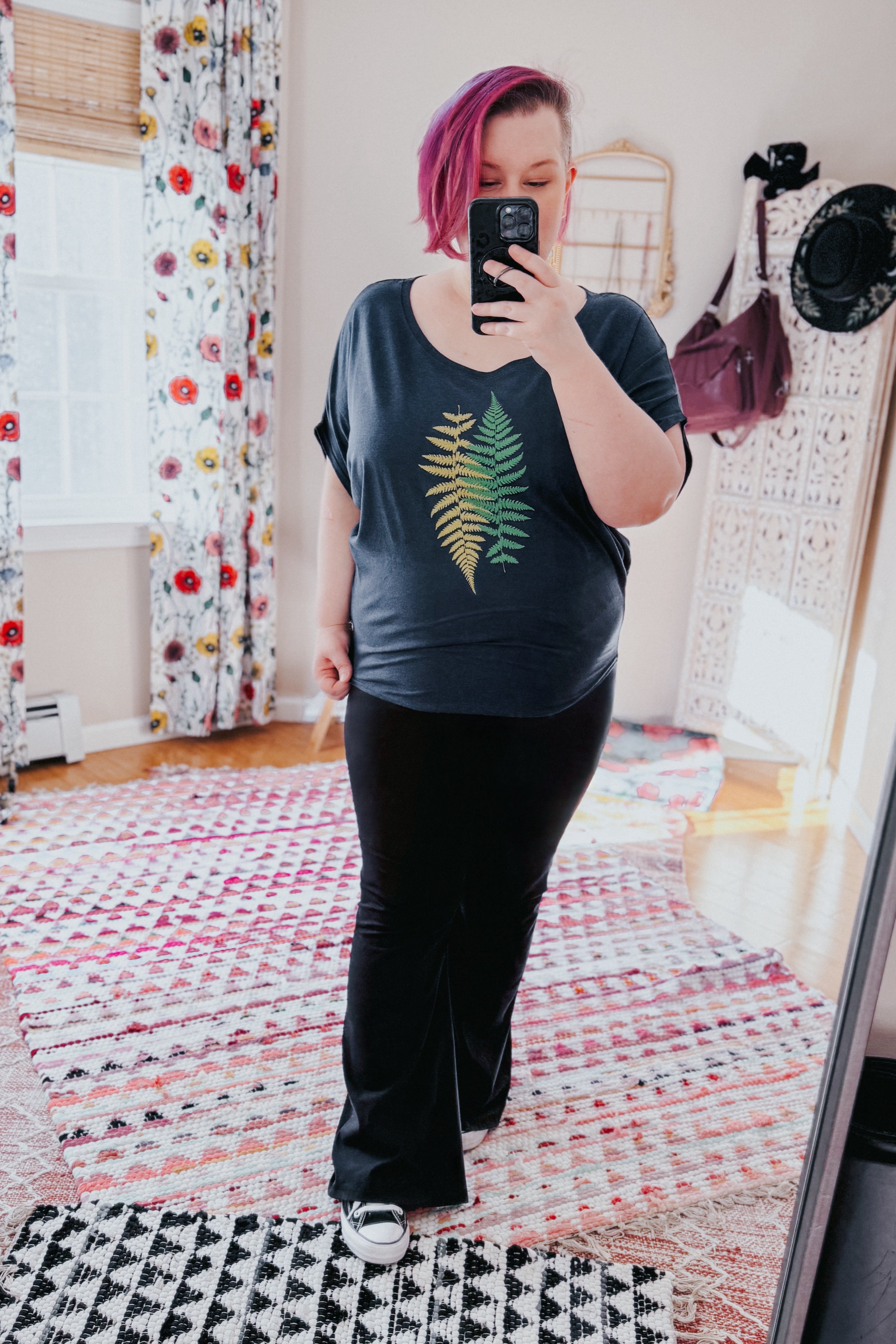 Woman with bright pink hair wearing navy bamboo dolman top with yellow and green fern print, black pants, and sneakers indoors