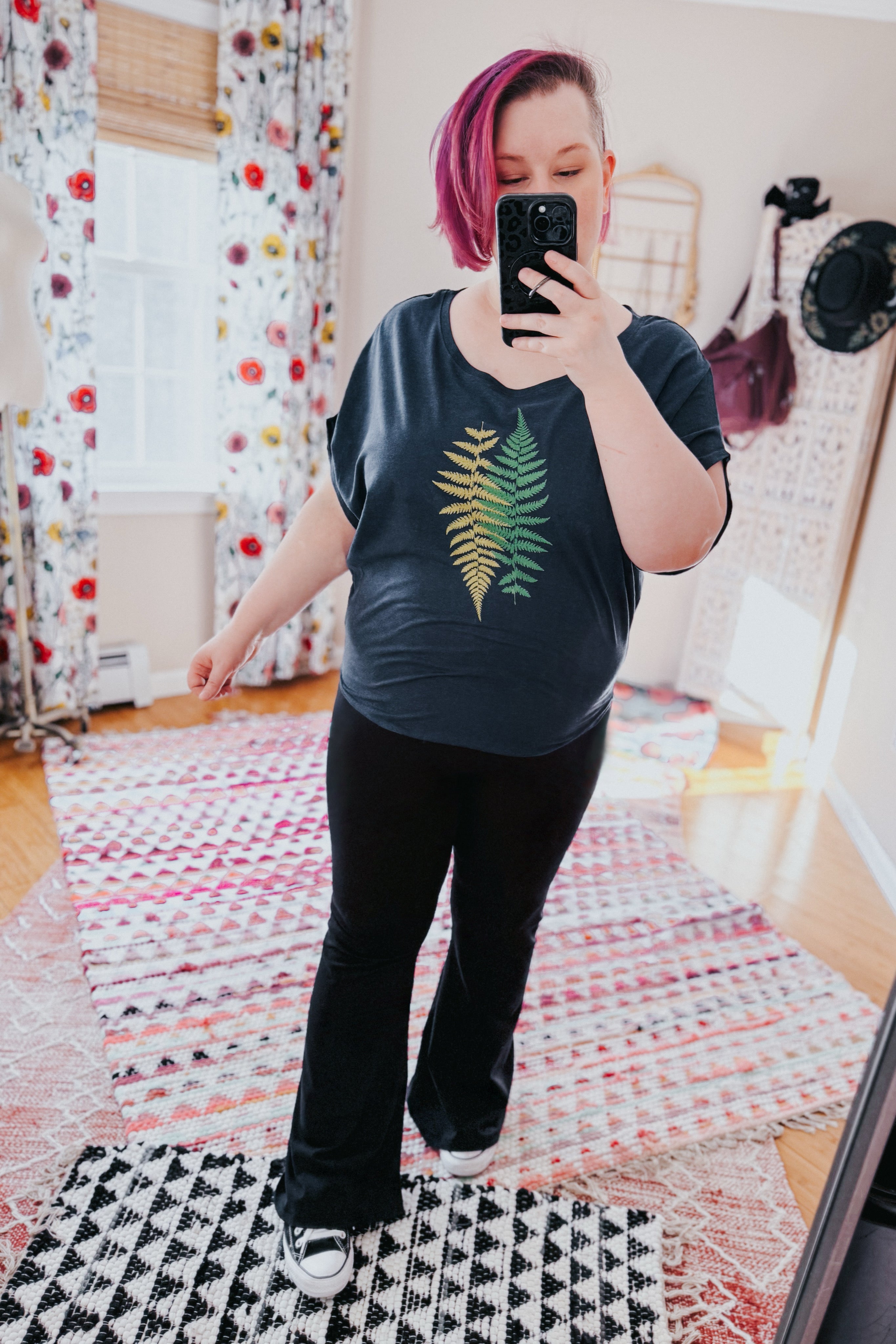 Woman with pink hair wearing navy bamboo dolman top with fern design and black pants taking mirror selfie in bright room