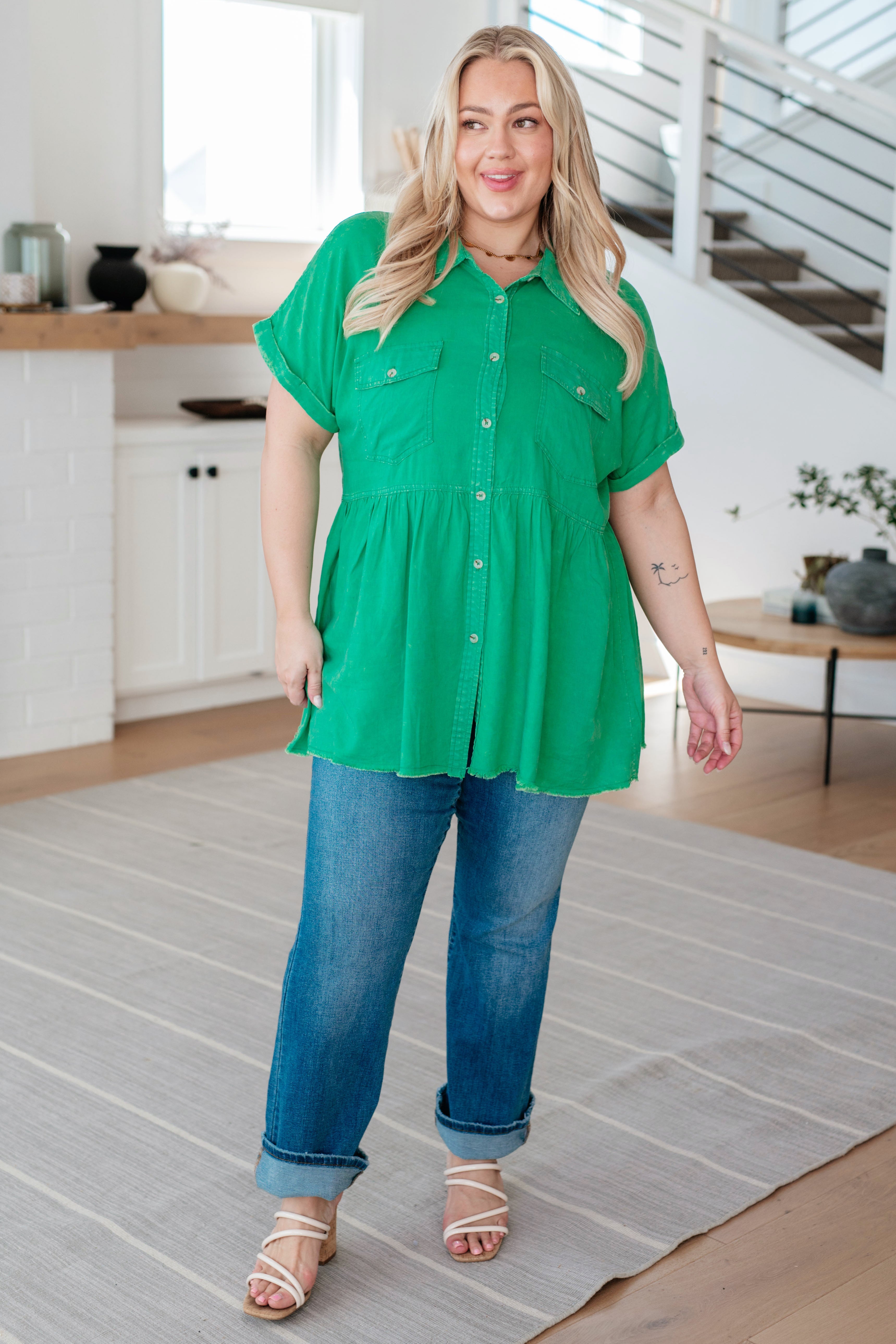 Woman wearing green button-up peplum blouse with rolled sleeves and relaxed blue jeans in bright modern home