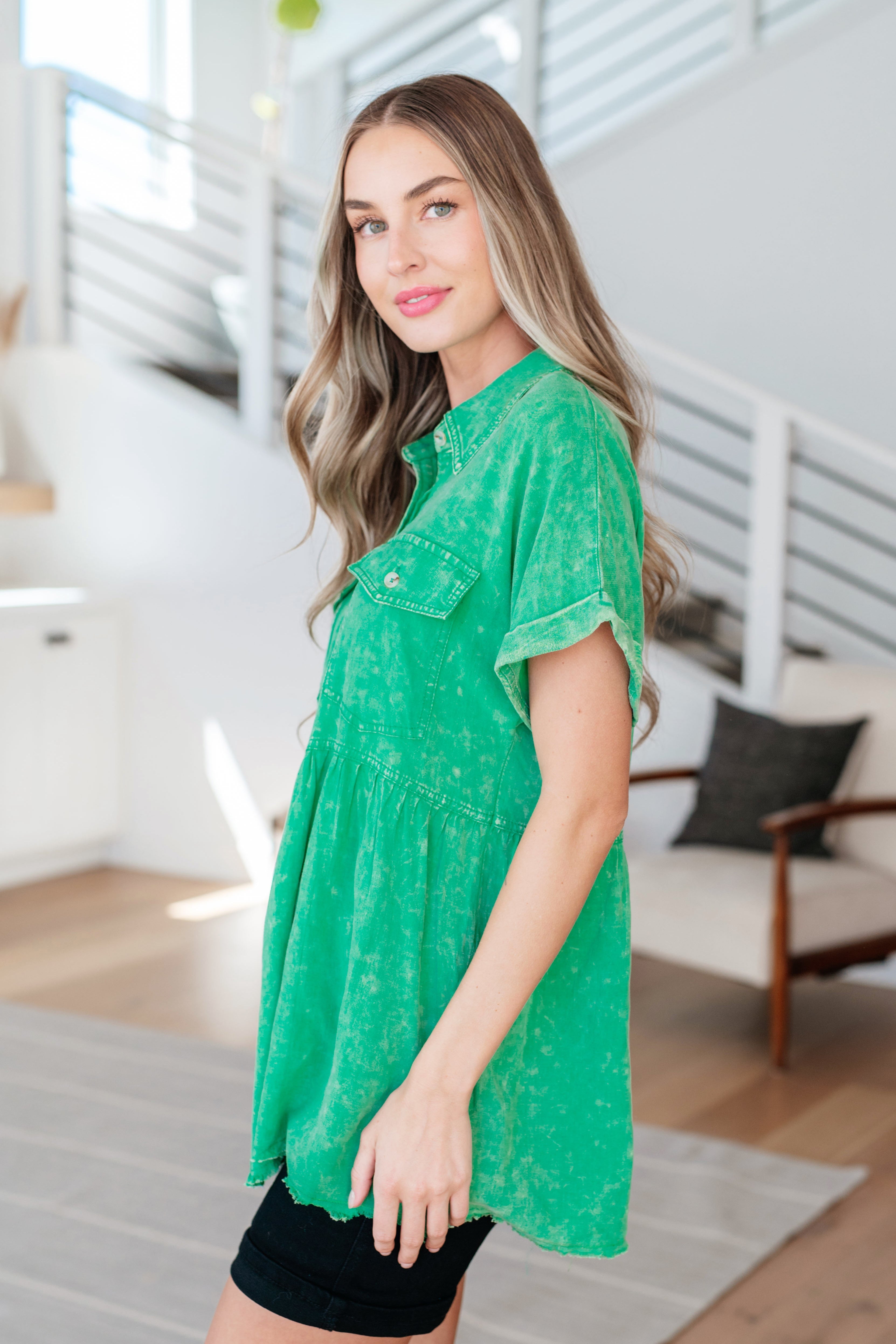 Woman wearing green button-up peplum blouse with rolled sleeves paired with black shorts in bright modern living room