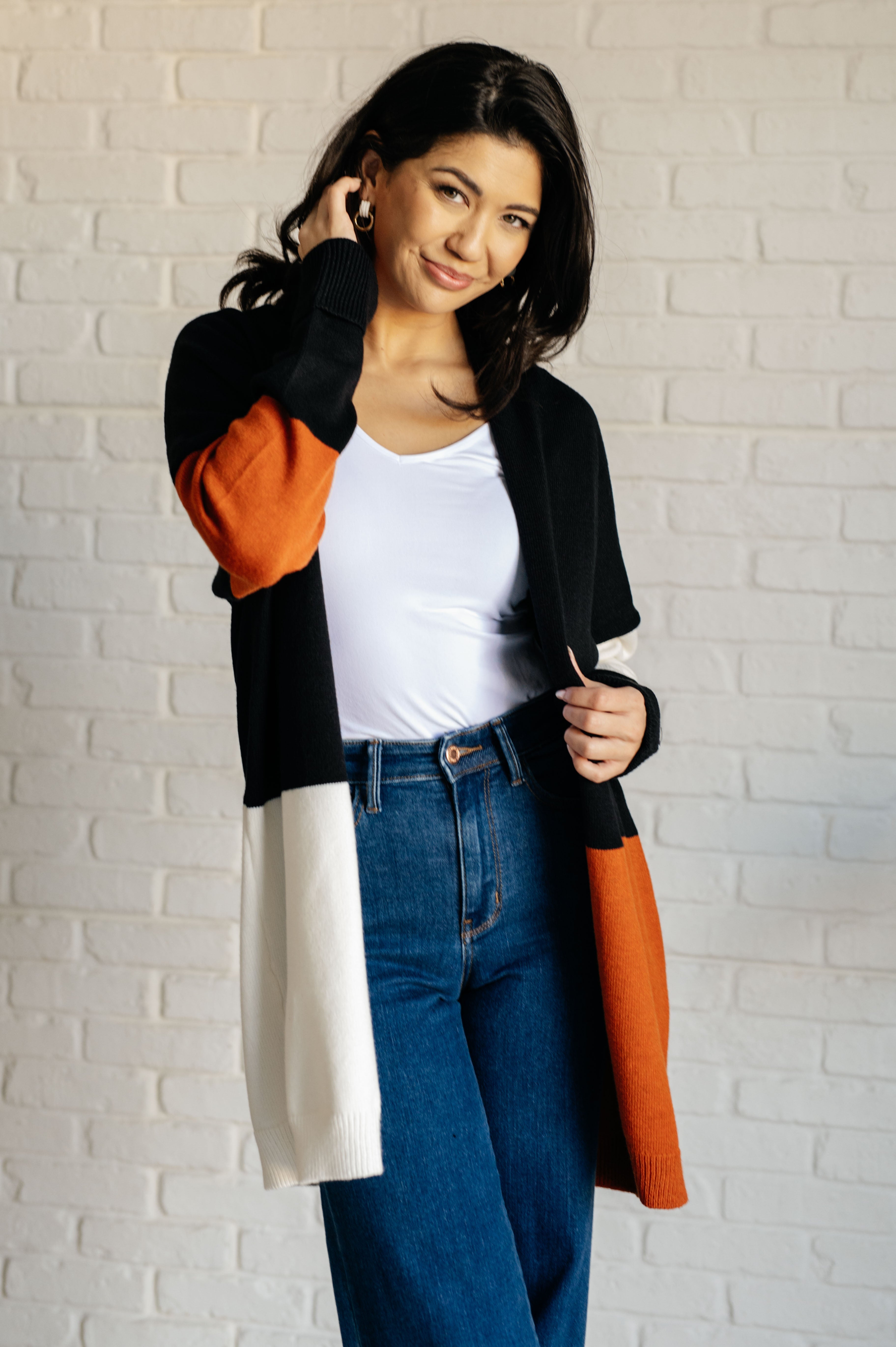 Woman wearing color block open front cardigan in black, orange, and white paired with white top and jeans indoors