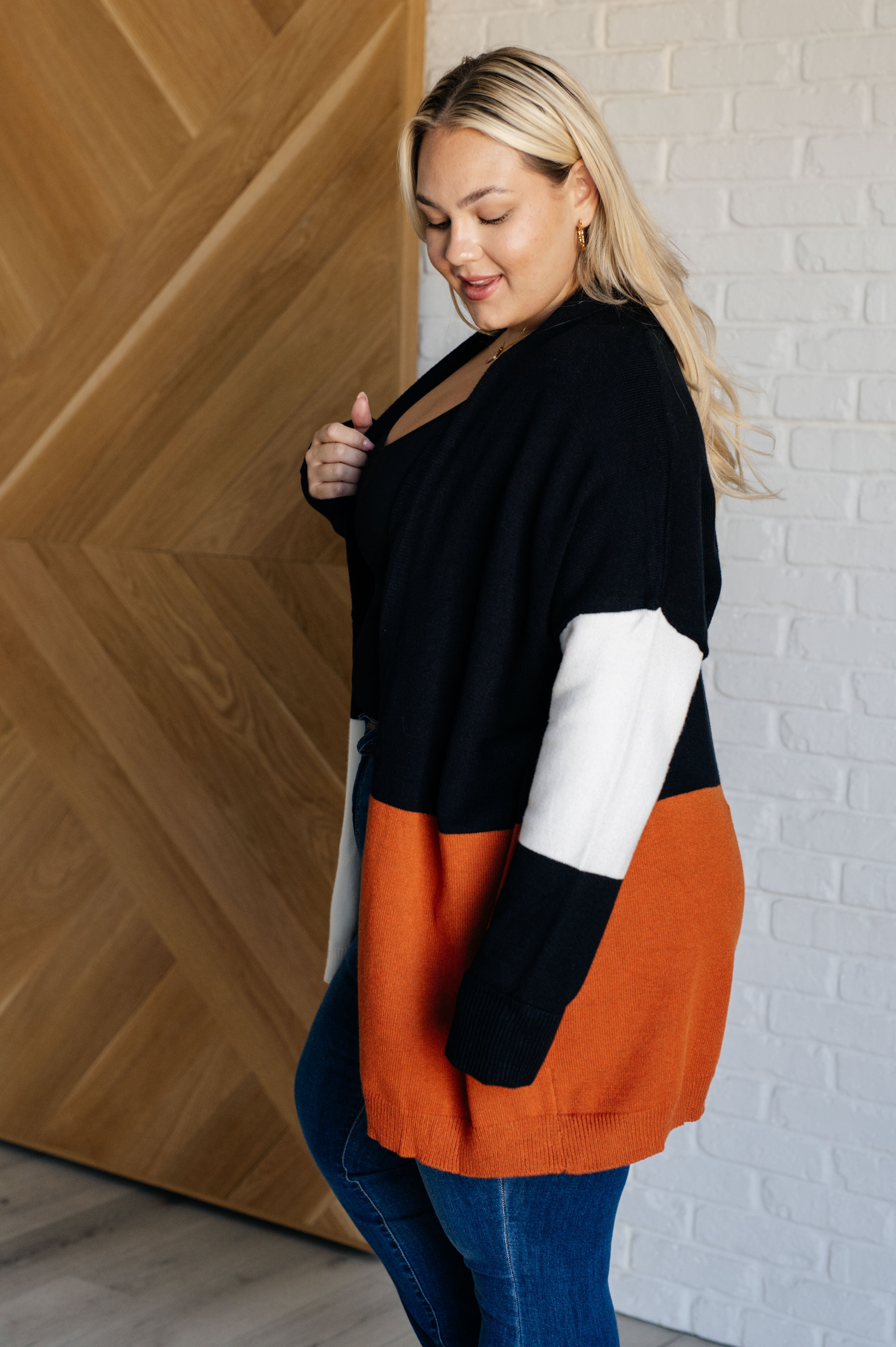 Curvy woman wearing black, white, and burnt orange color block cardigan over blue jeans in modern indoor setting
