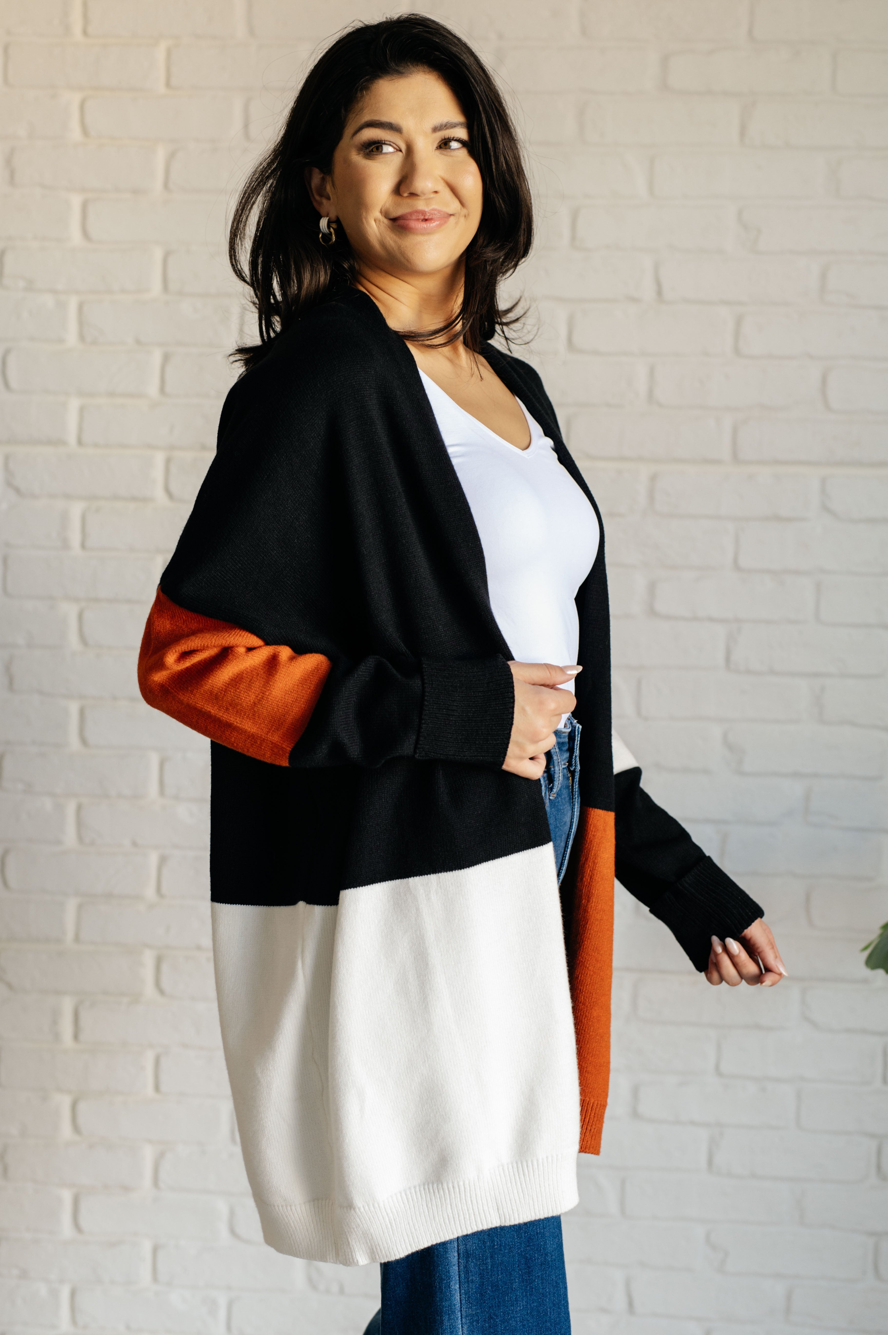 Woman wearing color block open front cardigan with black, white, and burnt orange panels over white top and jeans indoors