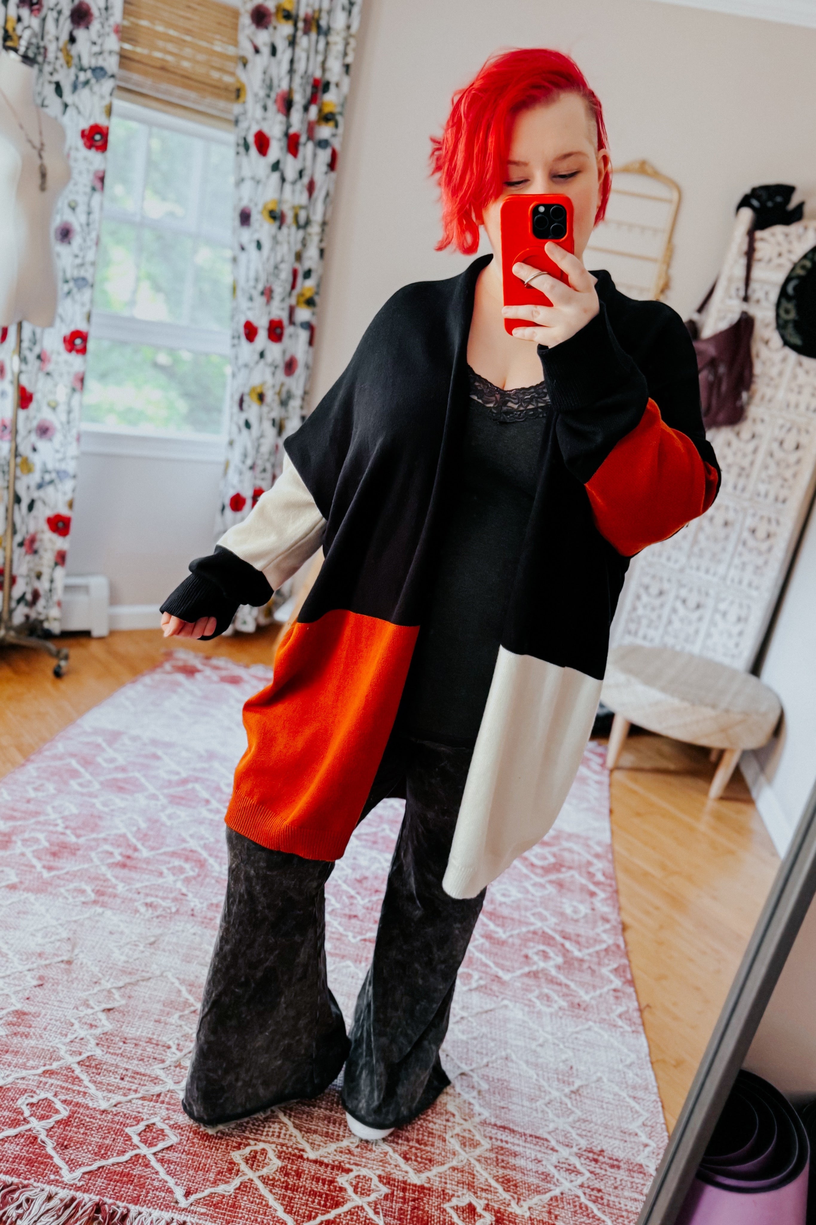 Woman with bright red hair wearing a color block black, orange, and cream open front cardigan taking mirror selfie indoors