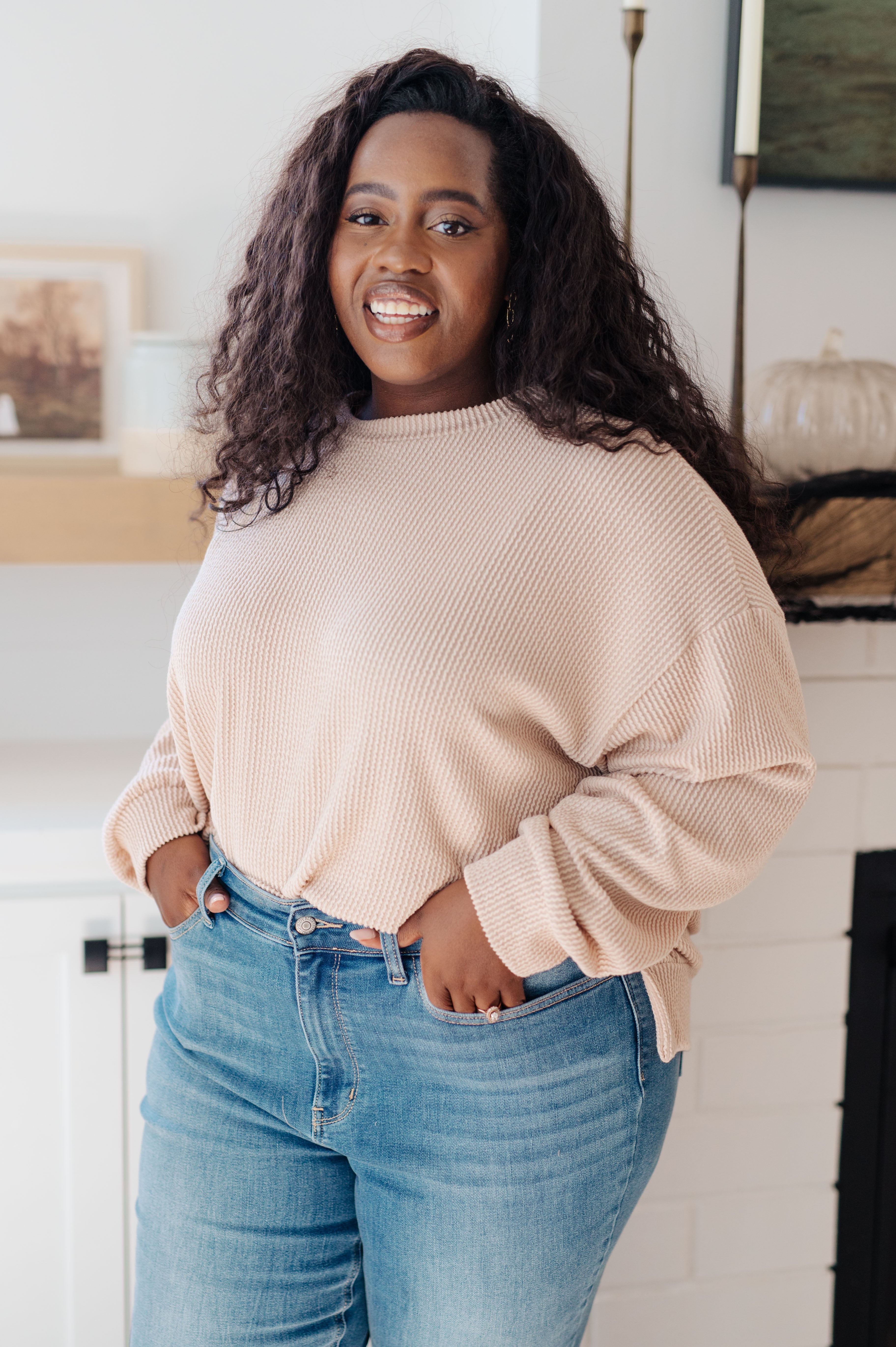 Smiling woman wearing beige textured pullover with relaxed fit and blue high-waisted jeans indoors