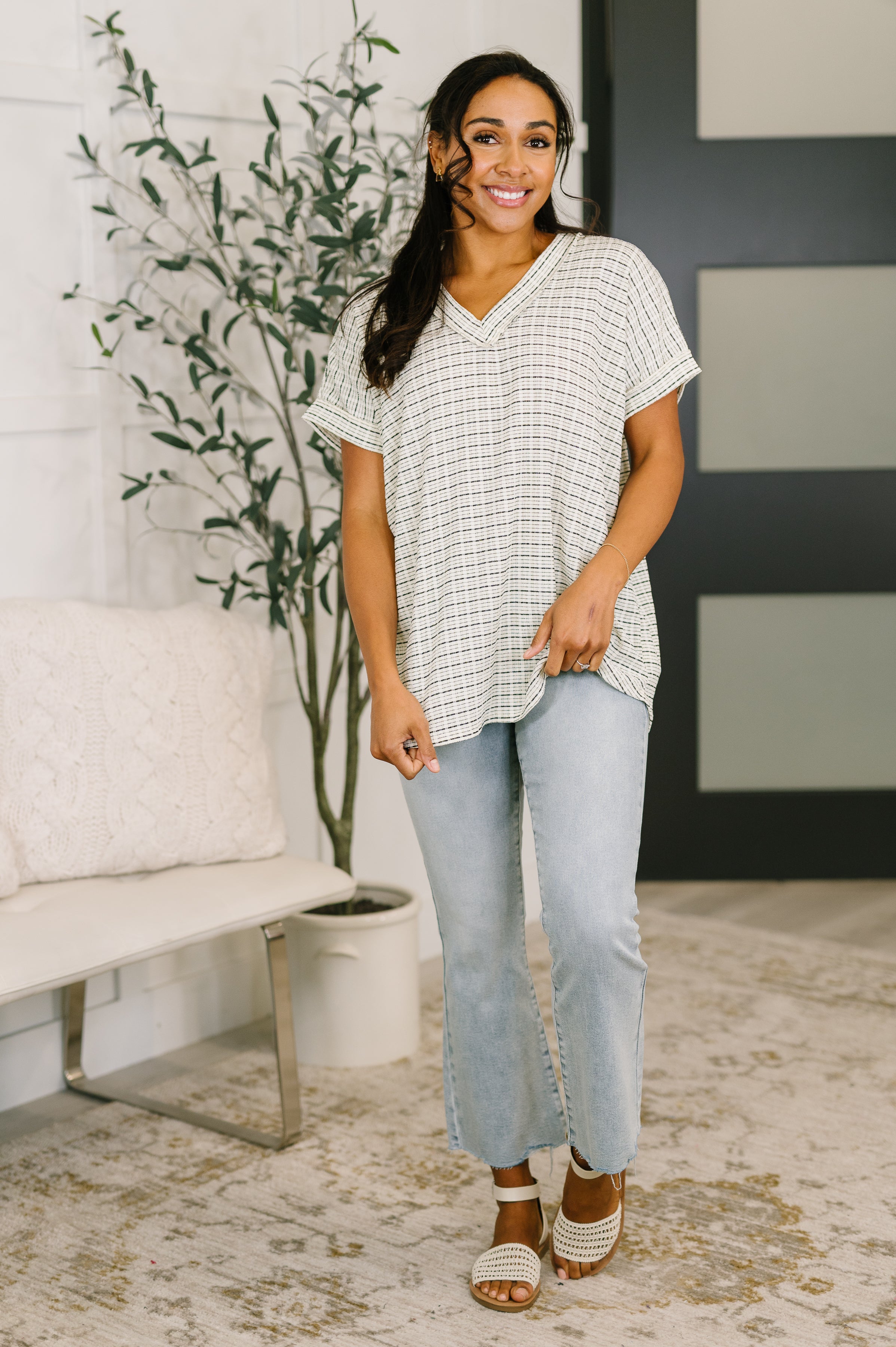 Smiling woman in a cream textured knit V-neck top paired with light wash jeans and woven sandals in a free-spirited style