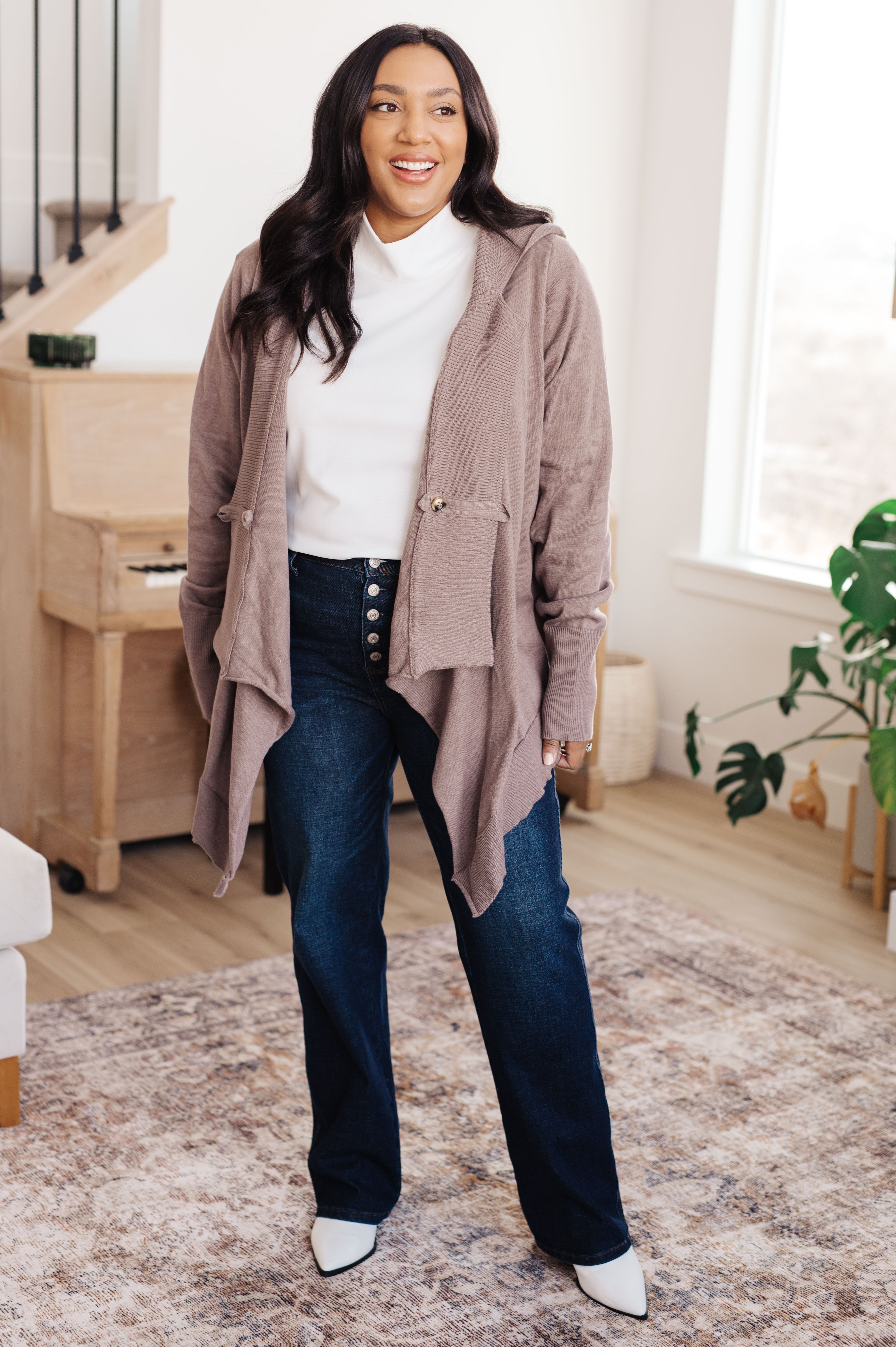 Woman wearing mocha-colored cozy cardigan with pockets, white top, dark high-waisted jeans, and white pointed shoes indoors