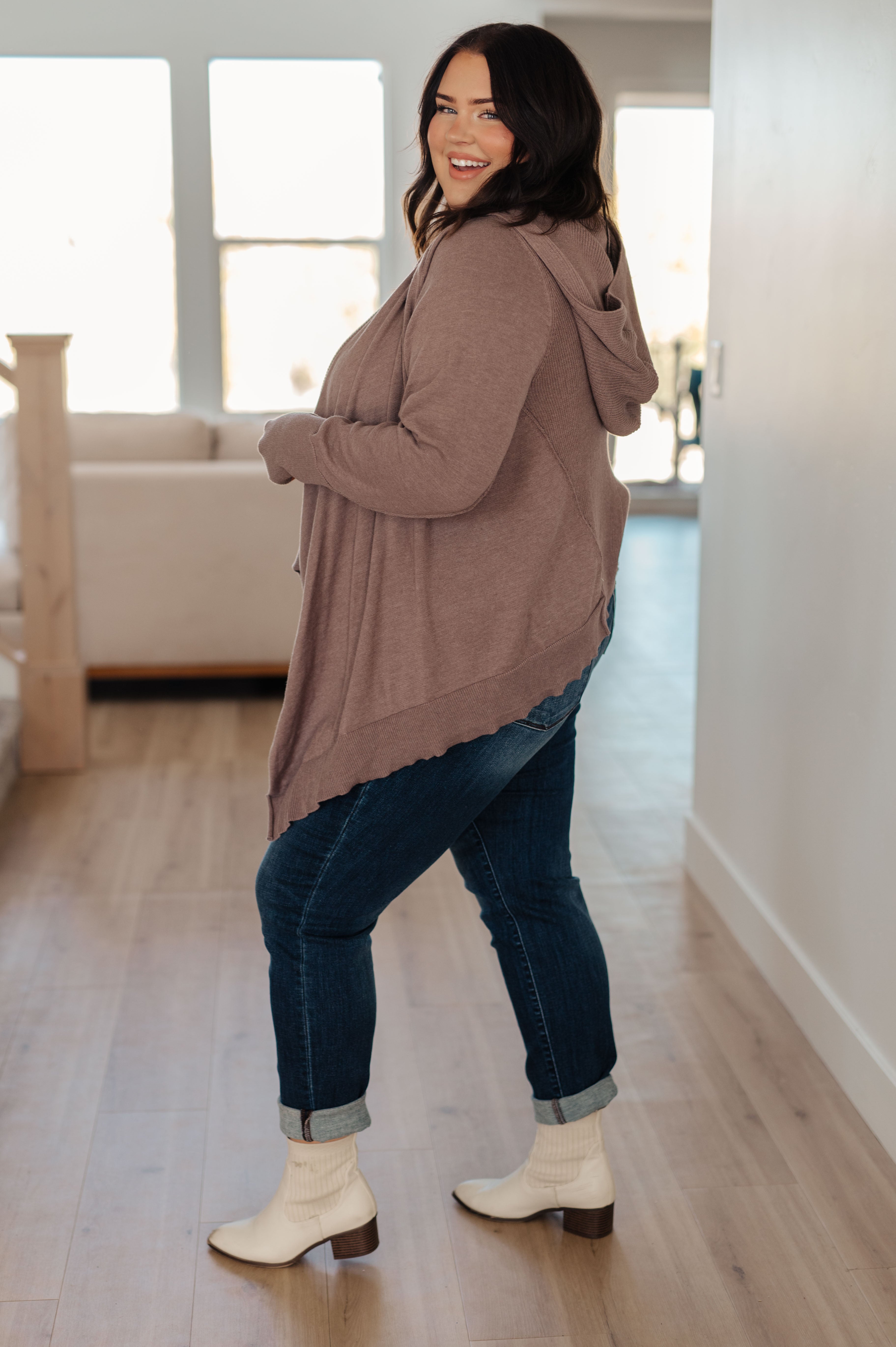 Smiling woman wearing mocha cozy cardigan with hood and cuffed dark jeans paired with cream ankle boots indoors