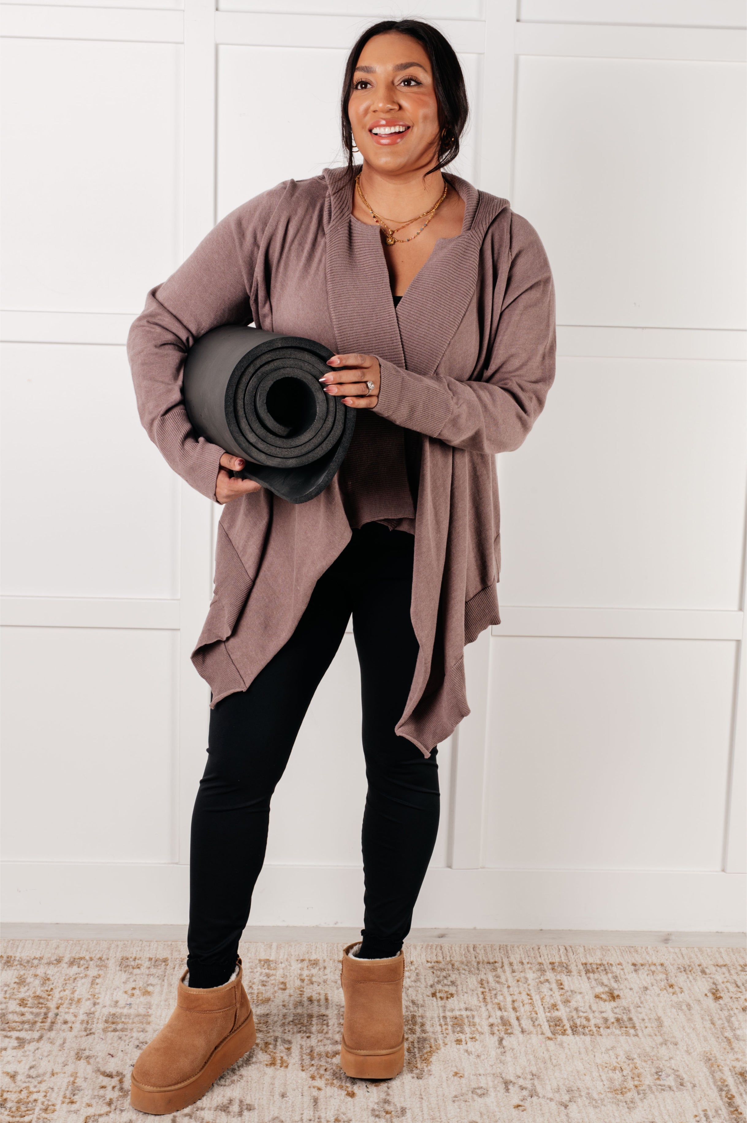 Woman wearing mocha draped cardigan layered over matching top holding yoga mat indoors