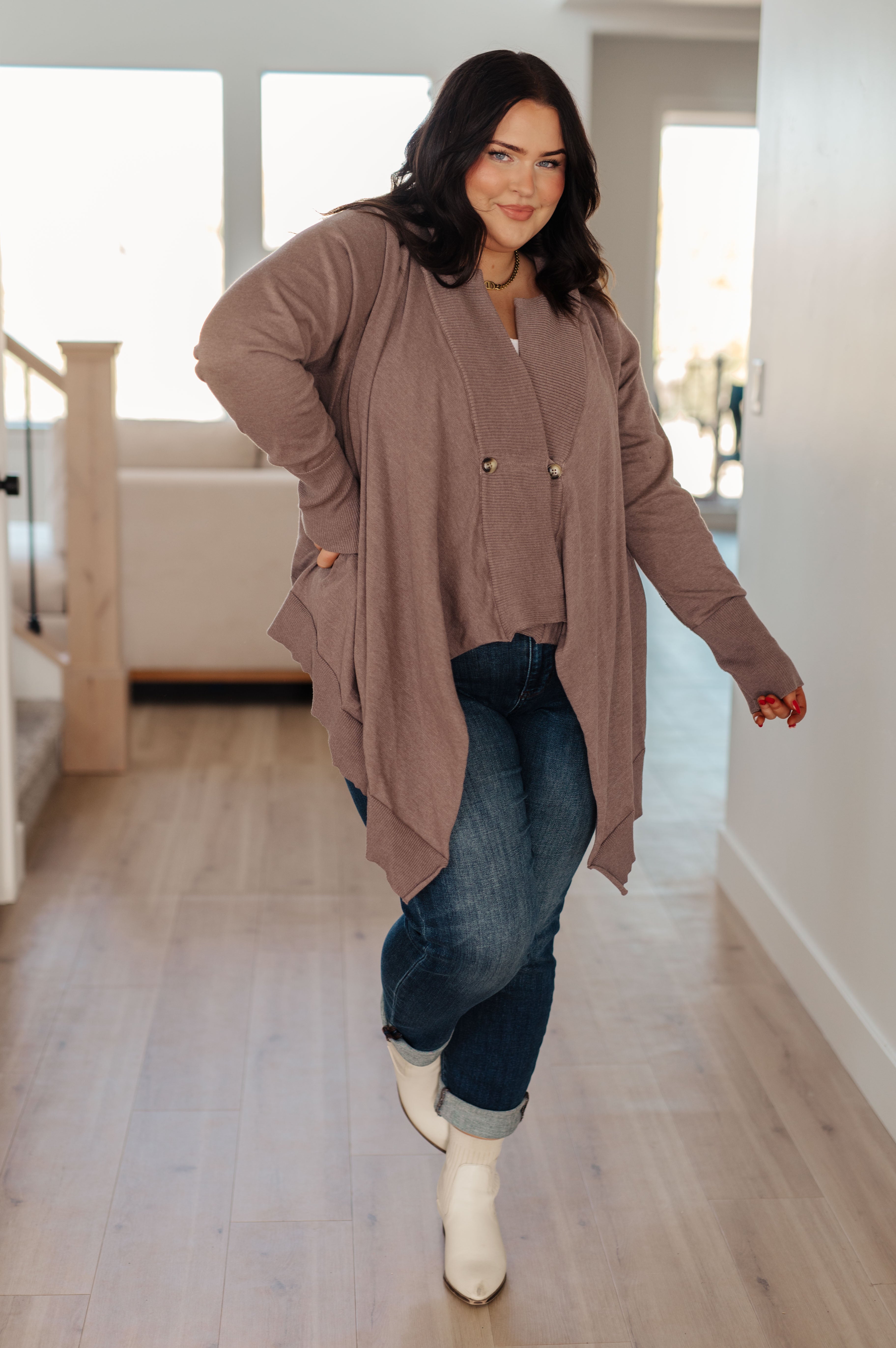 Plus-size woman indoors wearing a mocha draped cardigan with pockets styled with dark jeans and white boots
