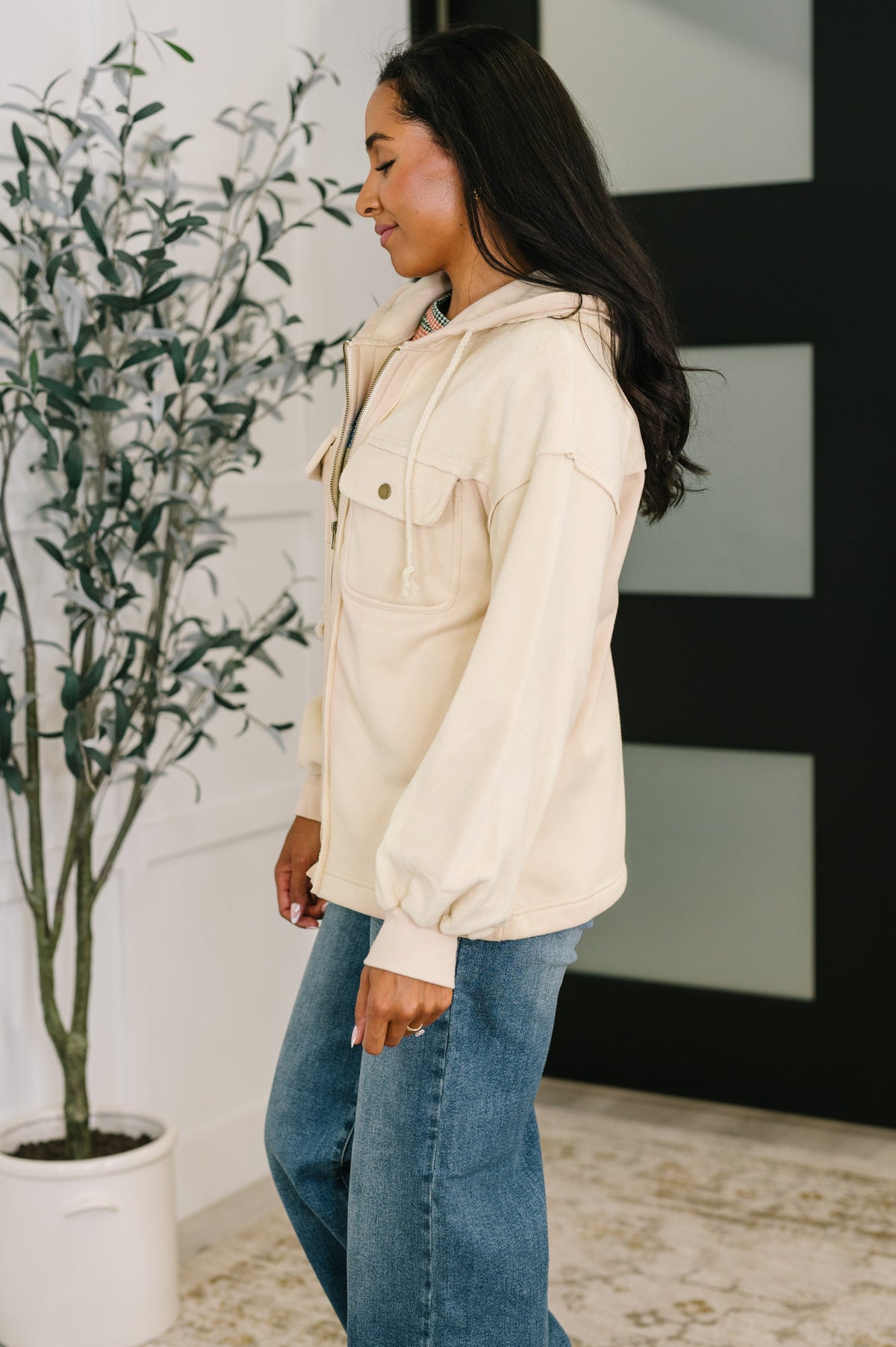 Woman in a beige zip front fleece hoodie with chest pockets and blue jeans standing beside a potted plant indoors