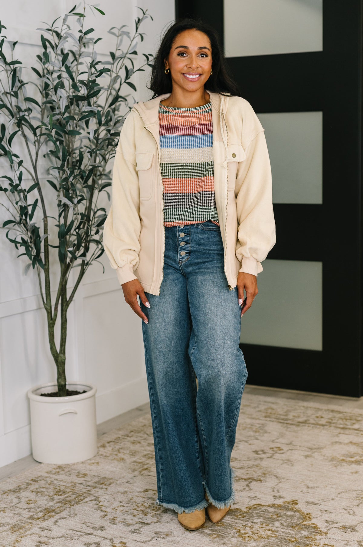 Woman wearing a beige zip front fleece jacket over a colorful striped top with wide leg jeans and tan shoes indoors