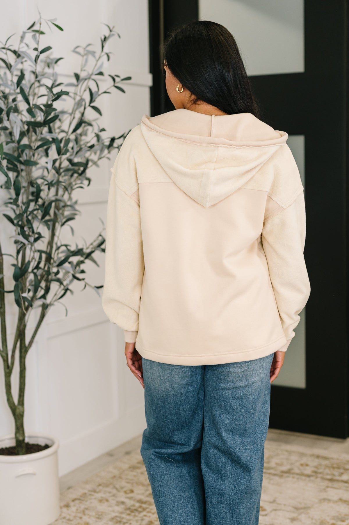 Woman seen from behind in a beige fleece hoodie and blue jeans standing beside a potted plant in a modern interior