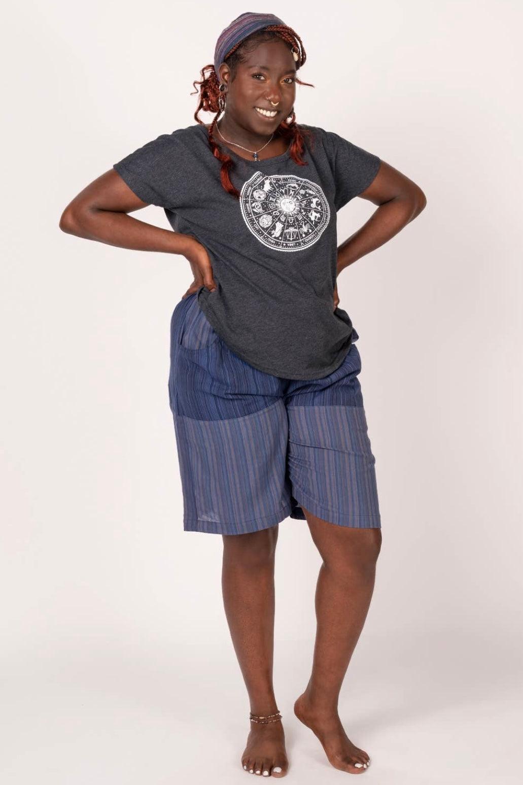 Smiling woman wearing a charcoal graphic tee with zodiac print and loose striped shorts standing barefoot
