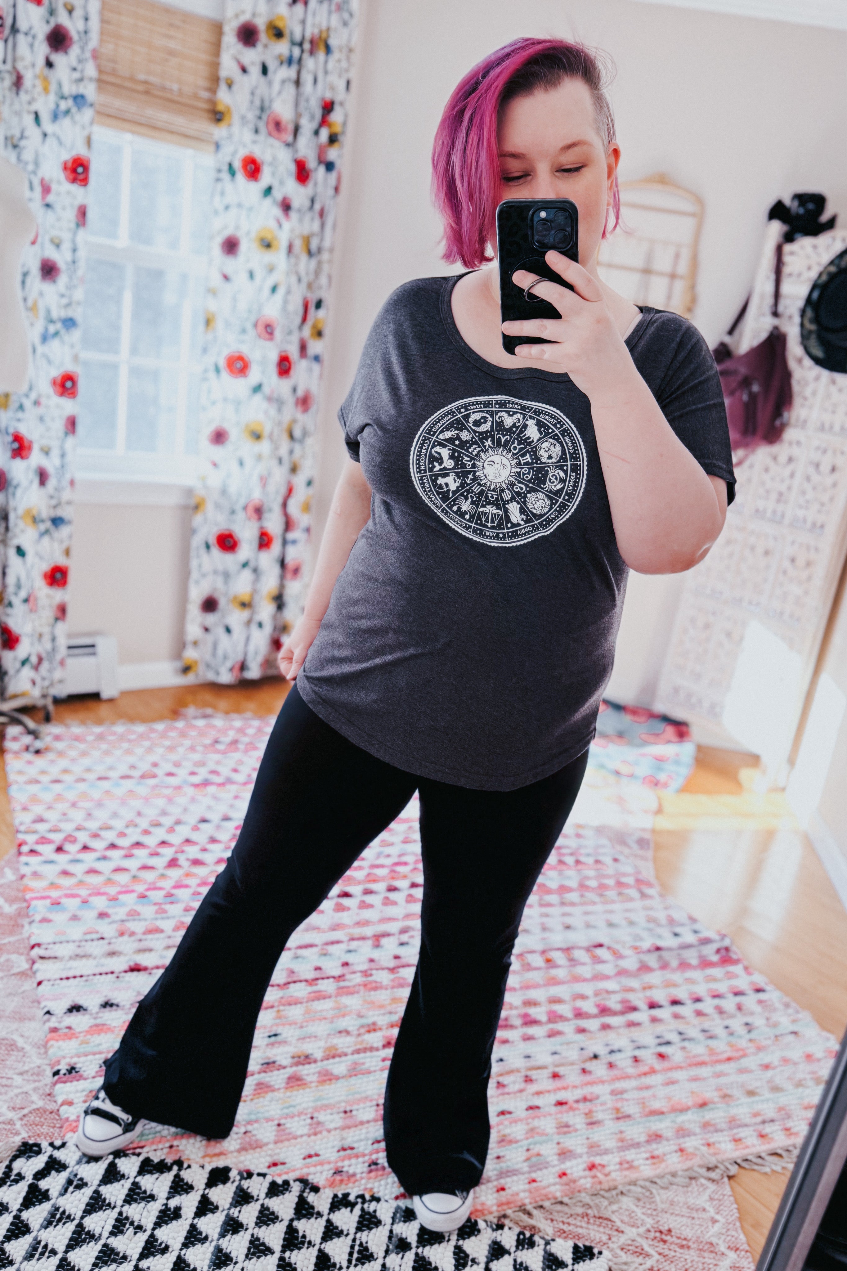 Woman wearing charcoal slouchy top with zodiac design paired with black flared pants taking mirror selfie indoors