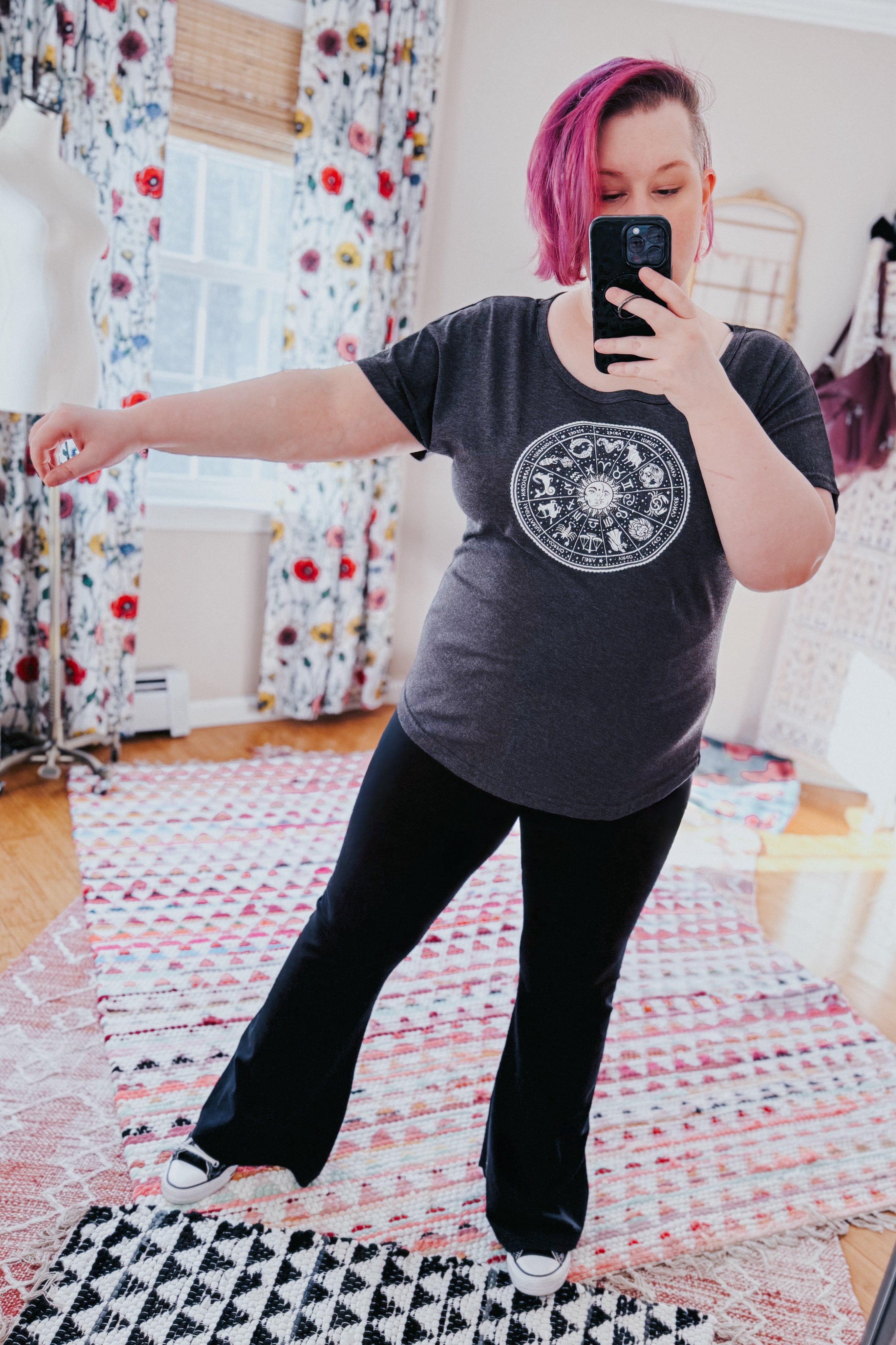 Woman with pink hair taking mirror selfie wearing charcoal zodiac circle top and black flared pants indoors